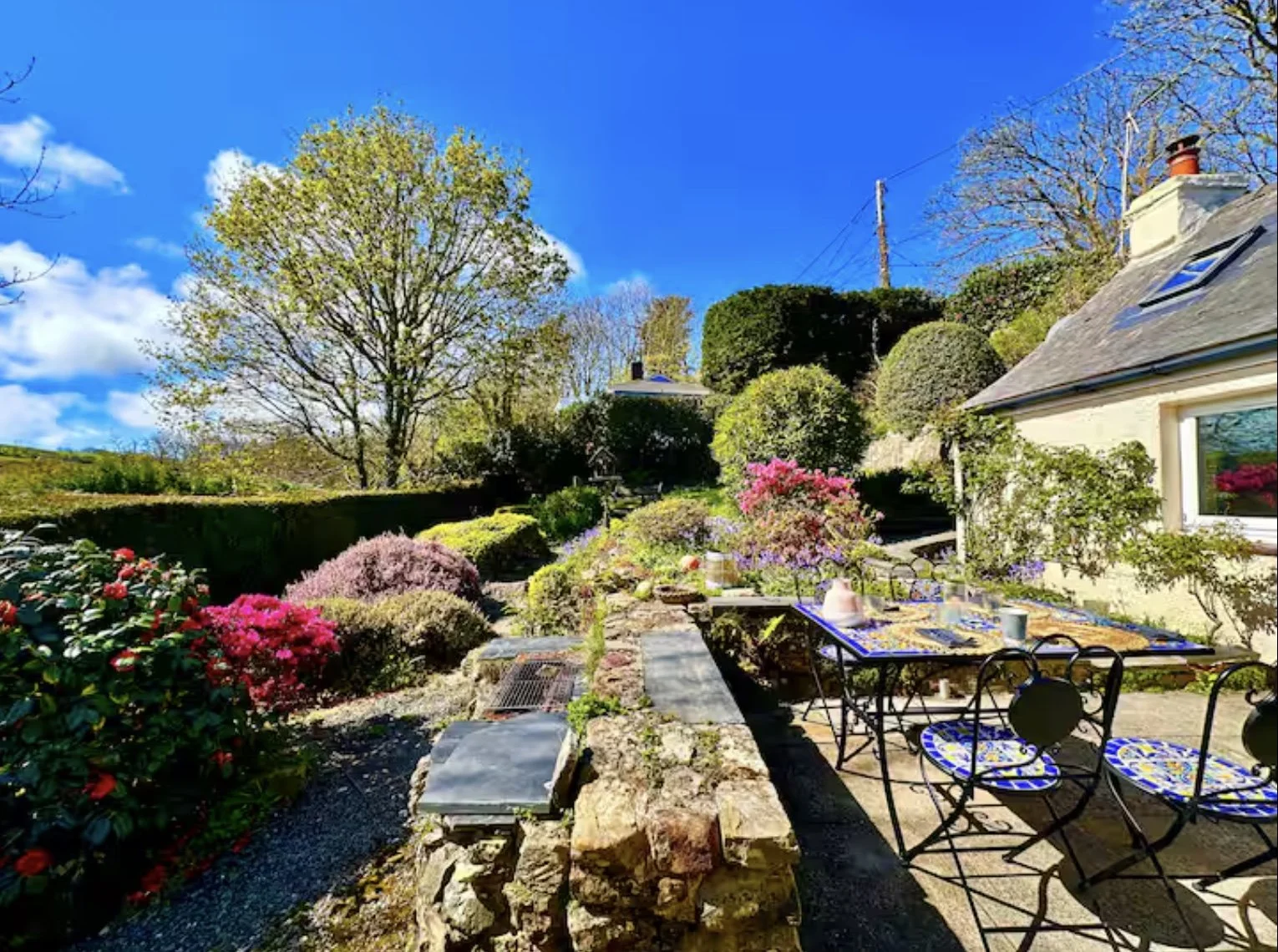 The Leap's colorful garden patio with a table sits in a garden surrounded by blooming bushes and trees, overlooking Little Haven's tranquil wooded valley.