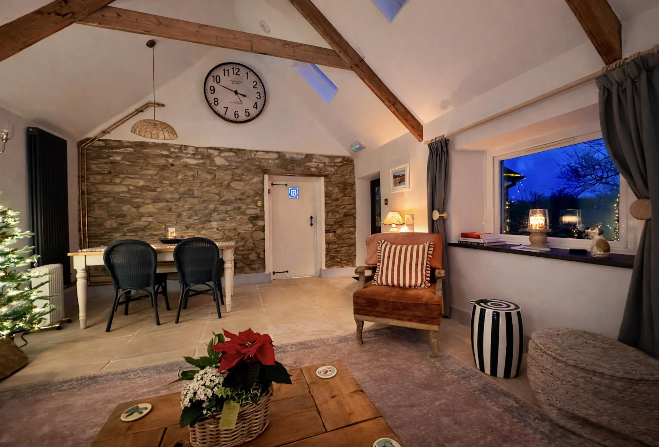 A cozy living room decorated for Christmas with a small Christmas tree, a wooden coffee table with a poinsettia, a brown armchair with a striped pillow, and a window showing a dark blue night sky with trees outside. There is a stone accent wall, a ro