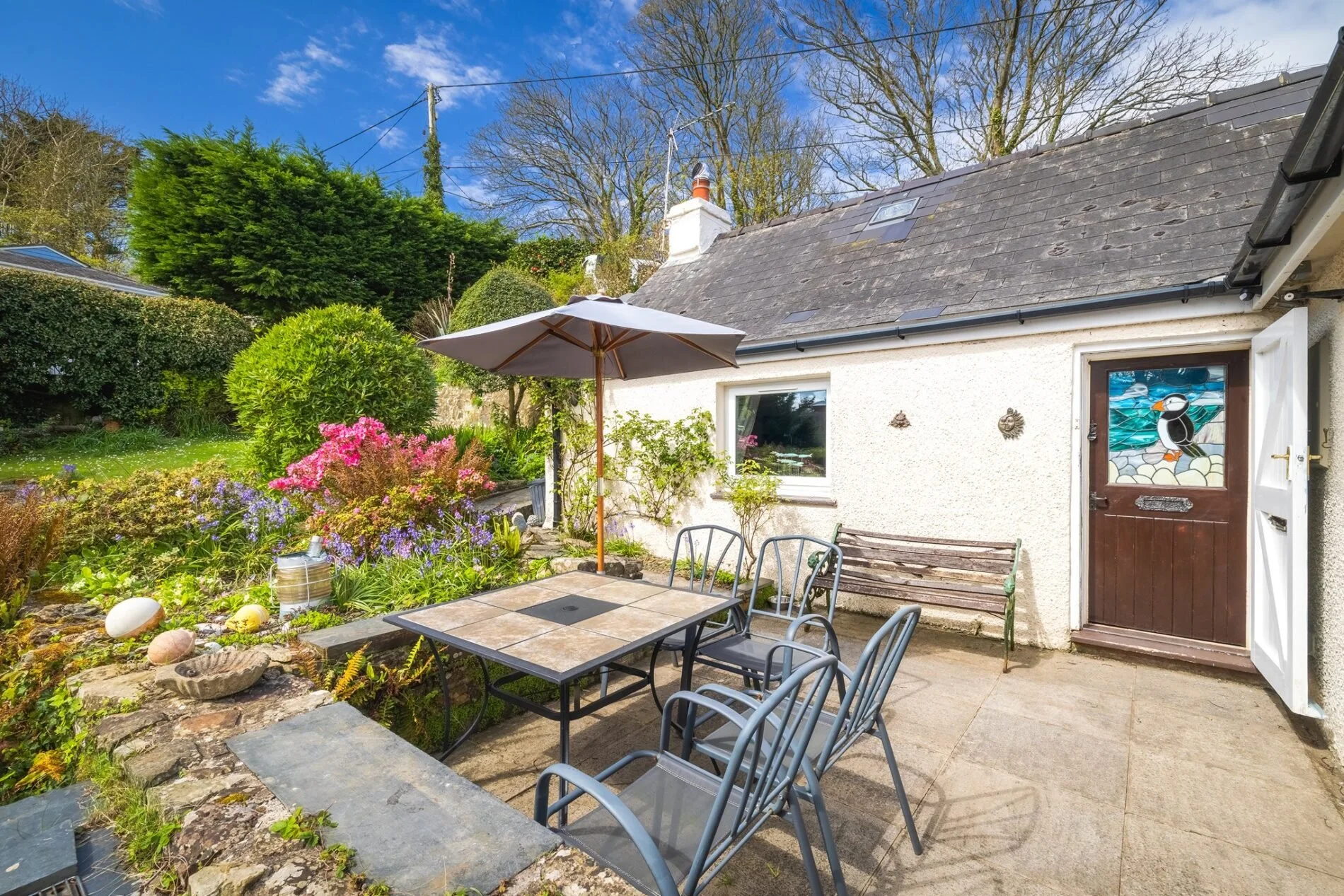 Backyard patio with a table, chairs, umbrella, and garden plants next to a white house with a window and door with a bird stain.