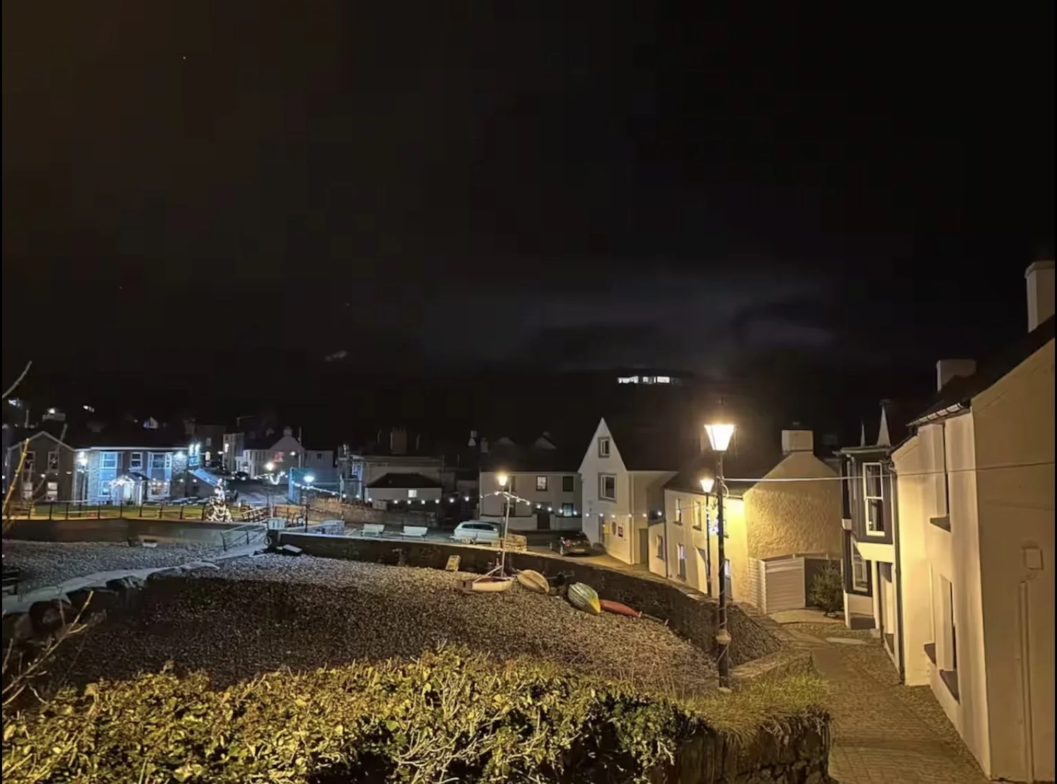 Nighttime view of a seaside village with lit street lamps, boats on a rocky shore, and houses with uniformly colored facades.