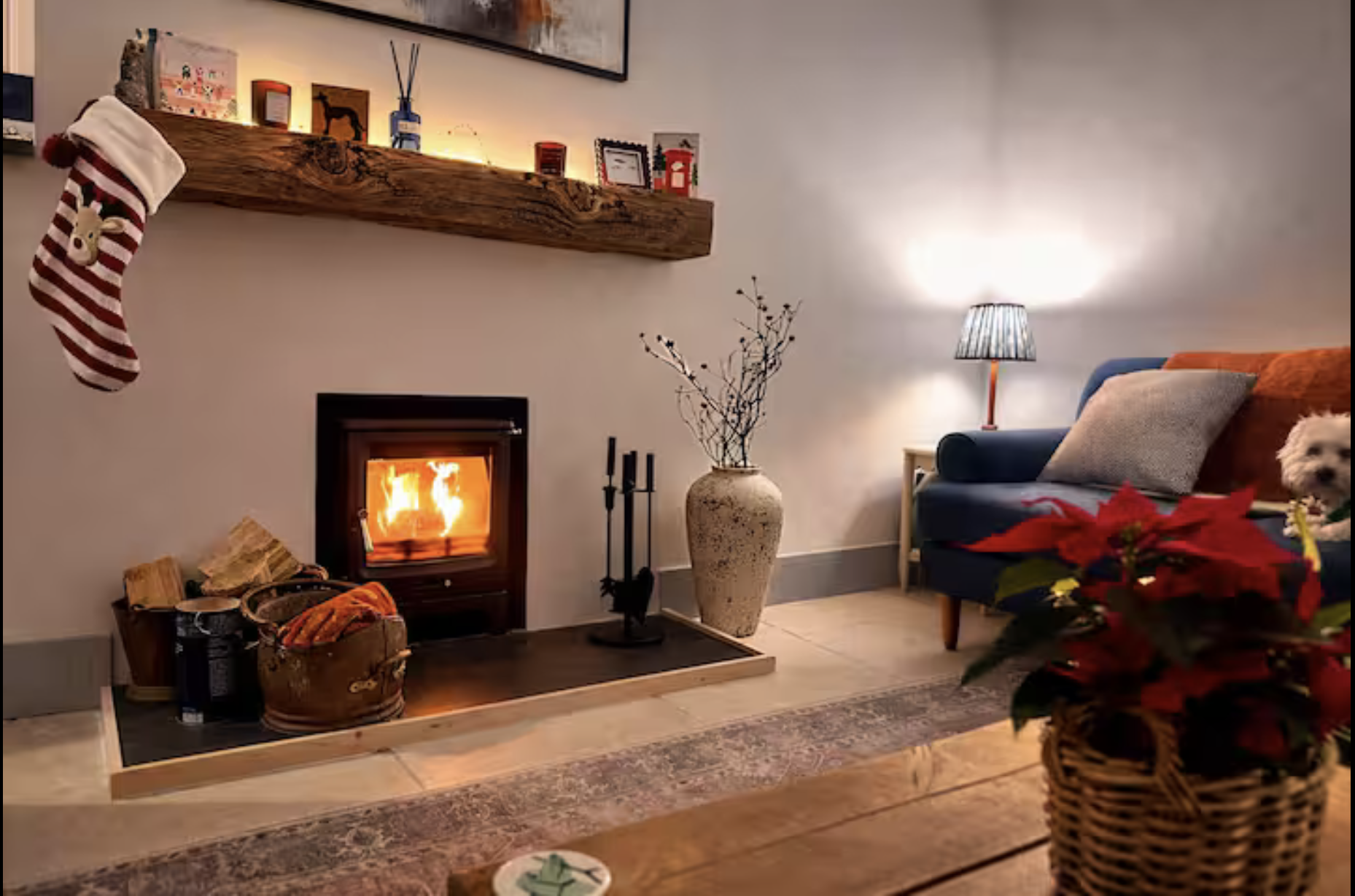 The Leap has a cosy living room and fireplace, a wooden mantel decorated with holiday pictures, a large vase with branches, a side table with a lamp, a sofa with cushions, and a poinsettia plant in a wicker basket.