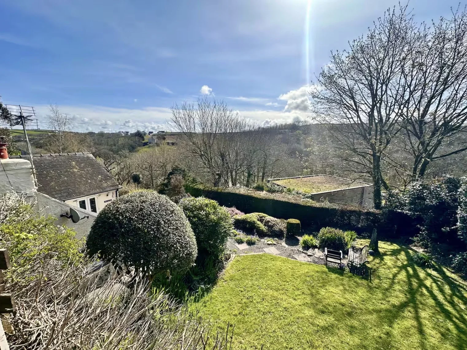 Sunshine over a garden with trimmed bushes, a group of chairs, and a large tree, with houses and hills in the background.