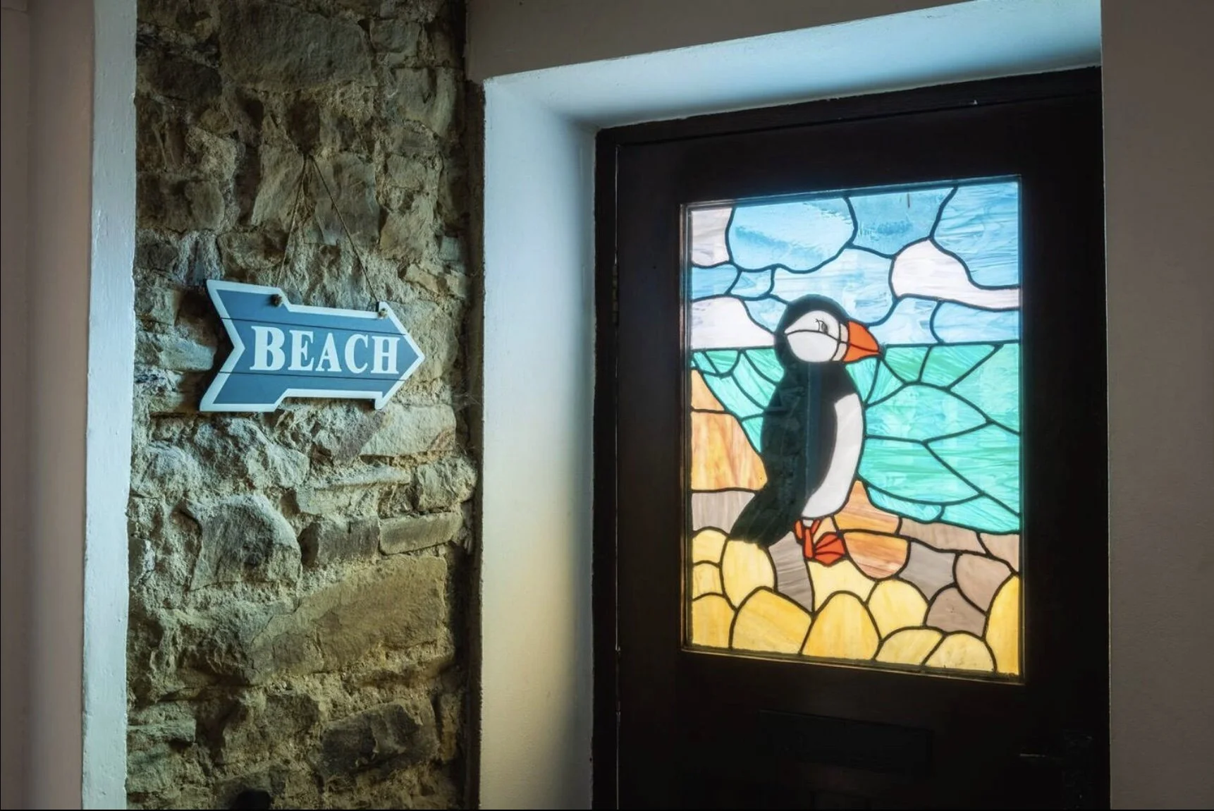 A decorative stained glass door featuring a puffin bird with a colorful background of blue sky, orange and yellow rocks. To the left, a blue arrow-shaped sign with the word 'BEACH' points towards the door, mounted on a stone wall.