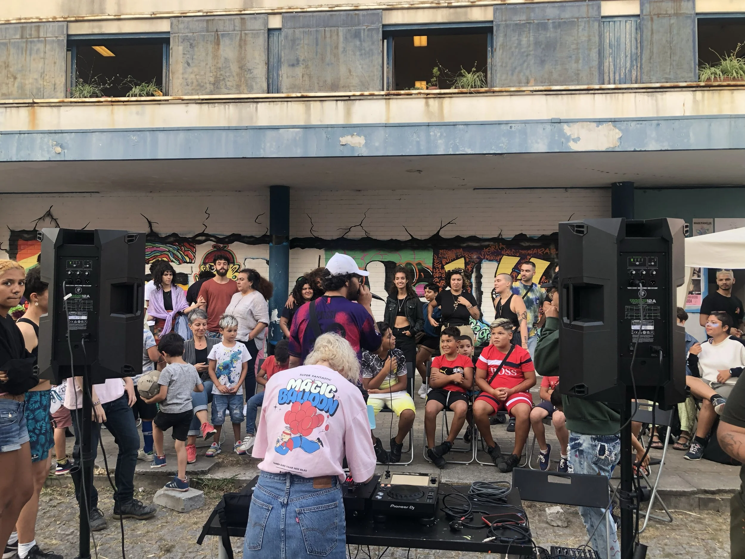 evento ao ar livre com pessoas, principalmente crianças, reunidas ao redor de um palco improvisado com caixas de som, em frente a um mural de grafite.