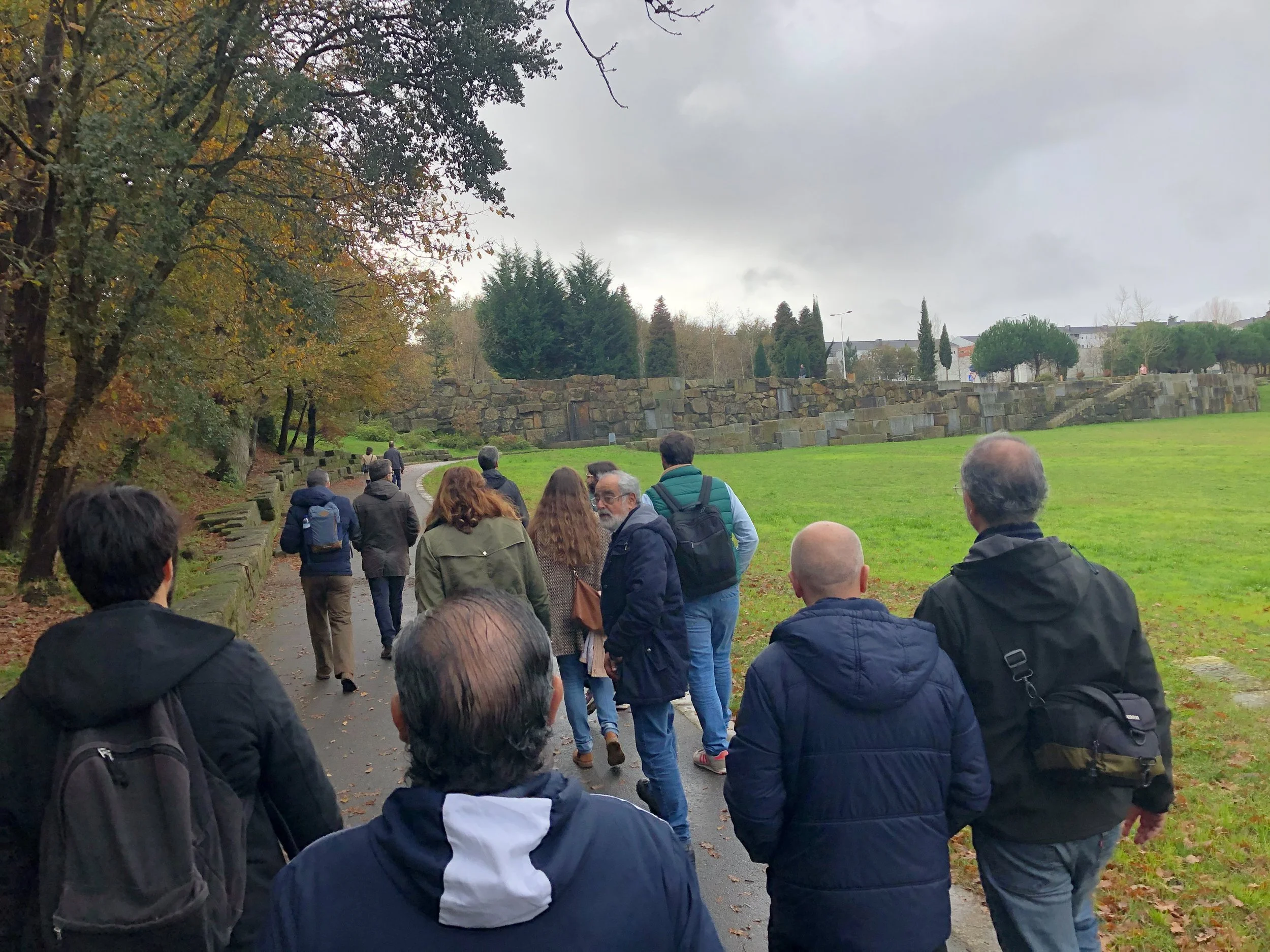 Grupo de pessoas caminhando por um caminho em um parque com árvores e uma parede de pedra ao fundo em um dia nublado.