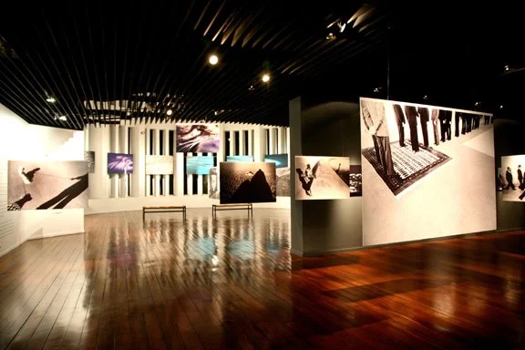 Exposição de fotografias em uma galeria de arte moderna com iluminação suave e piso de madeira.
