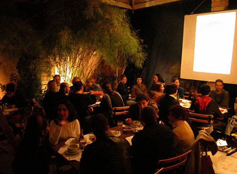 Pessoas assistindo a uma apresentação em um salão interno com iluminação amarelada e vegetação ao fundo.