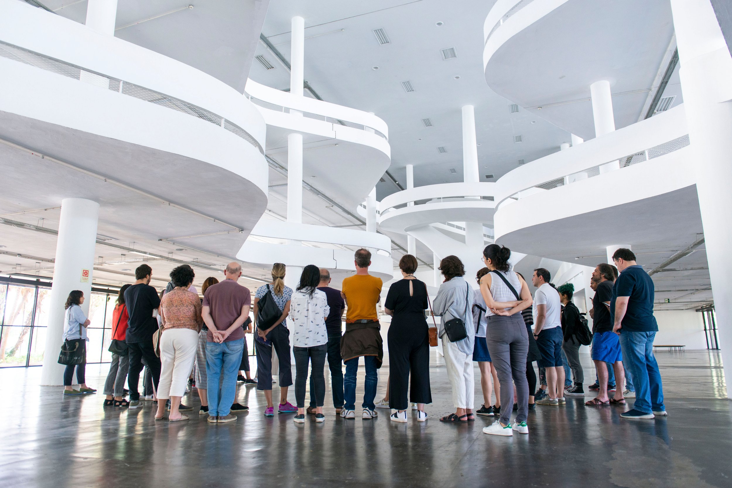 Grupo de pessoas assistindo uma apresentação em um espaço interno moderno com arquitetuta branca e escadas suspensas em espiral.