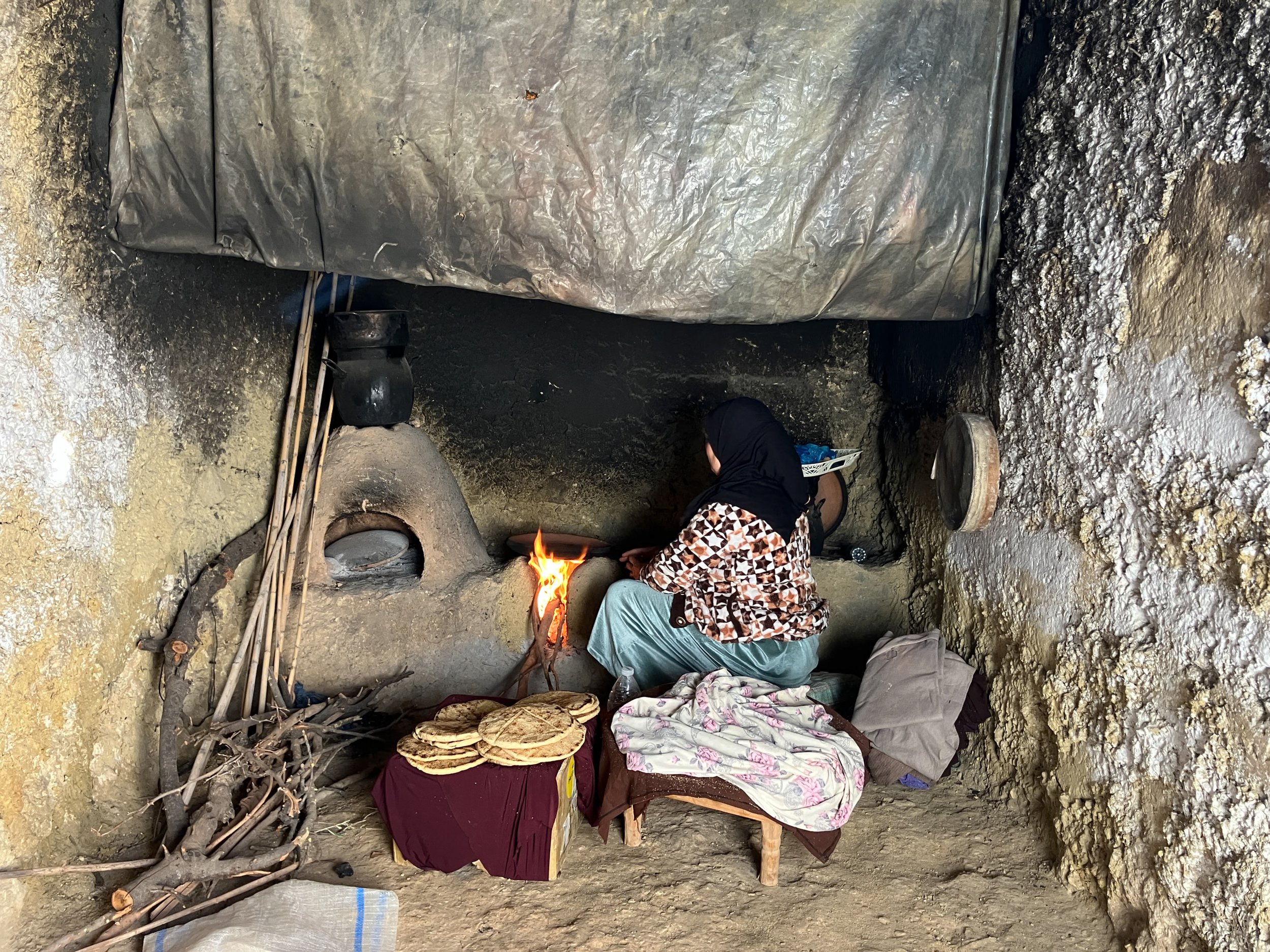 Pessoa sentada junto a uma fogueira em uma caverna rústica, com utensílios e roupas ao redor.