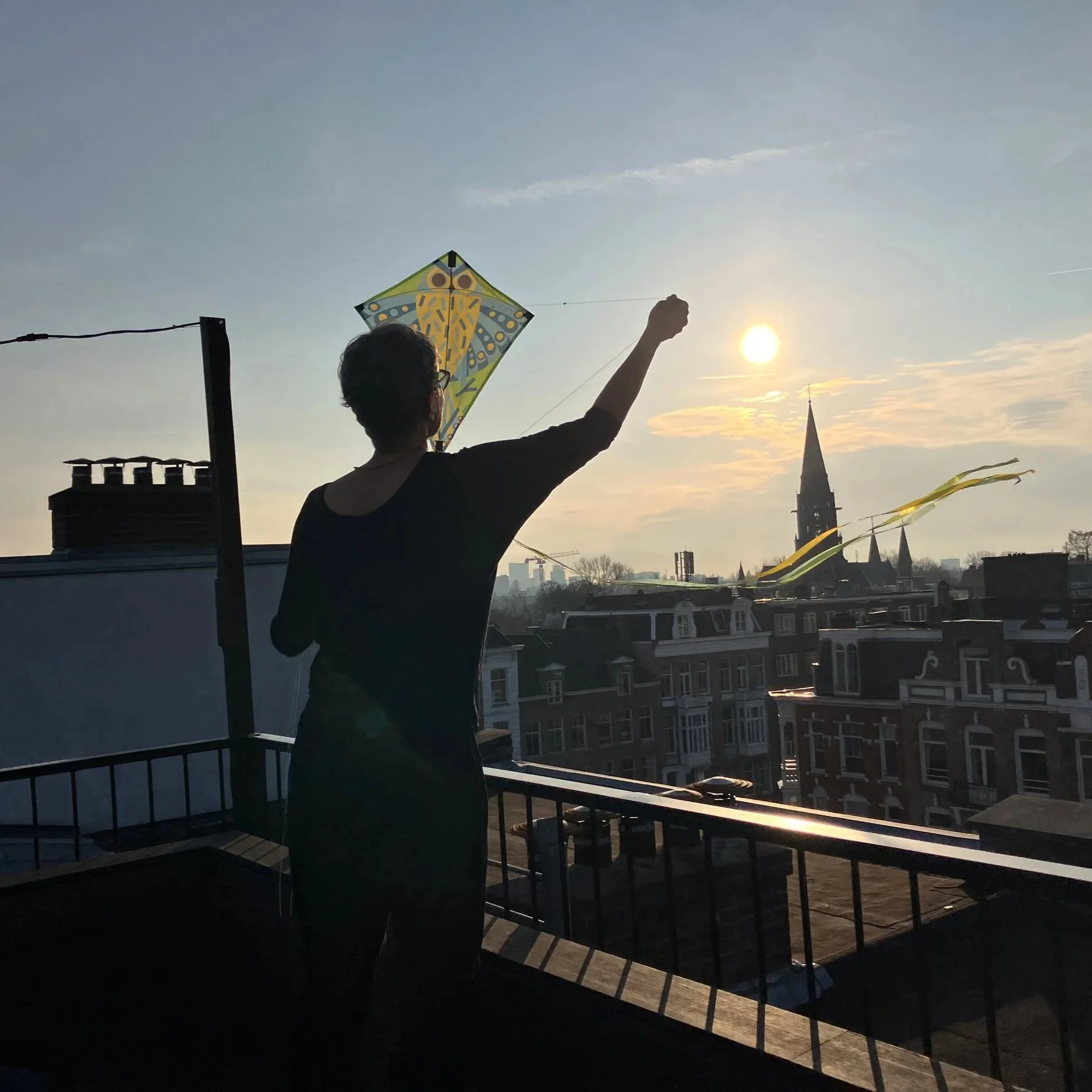 Pessoa de costas segurando uma pipa com o sol ao fundo em um terraço com vista para Igreja e casas na cidade.