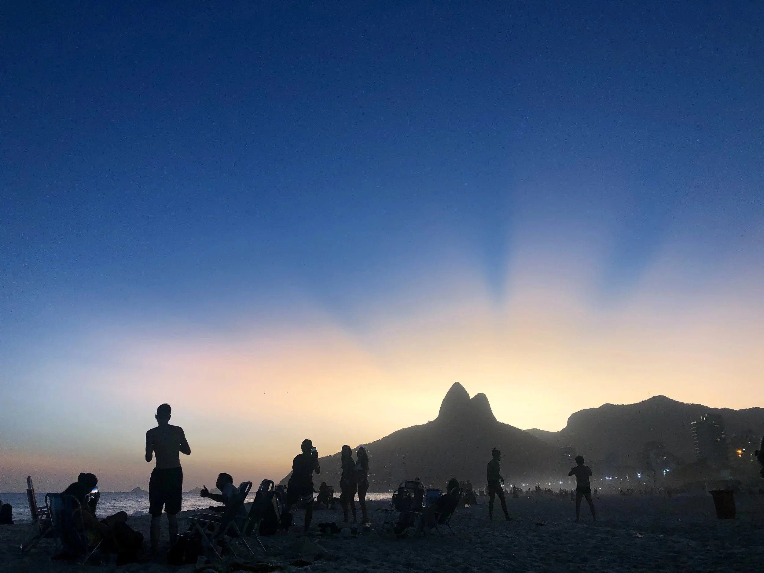 Pessoas na praia ao entardecer, com uma formação rochosa ao fundo, enquanto o céu passa do azul ao laranja.