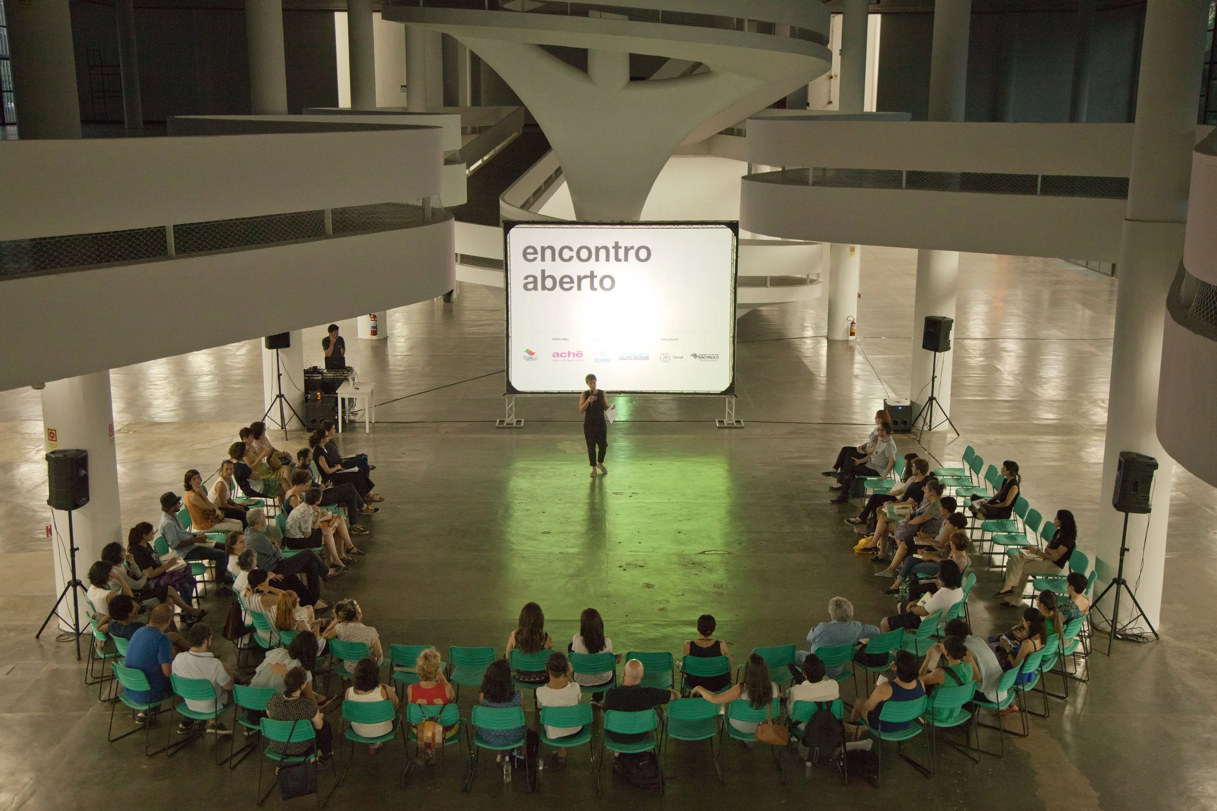 Evento em um espaço amplo com pessoas sentadas formando um círculo, assistindo uma apresentação com tela grande que diz 'encontro aberto'.