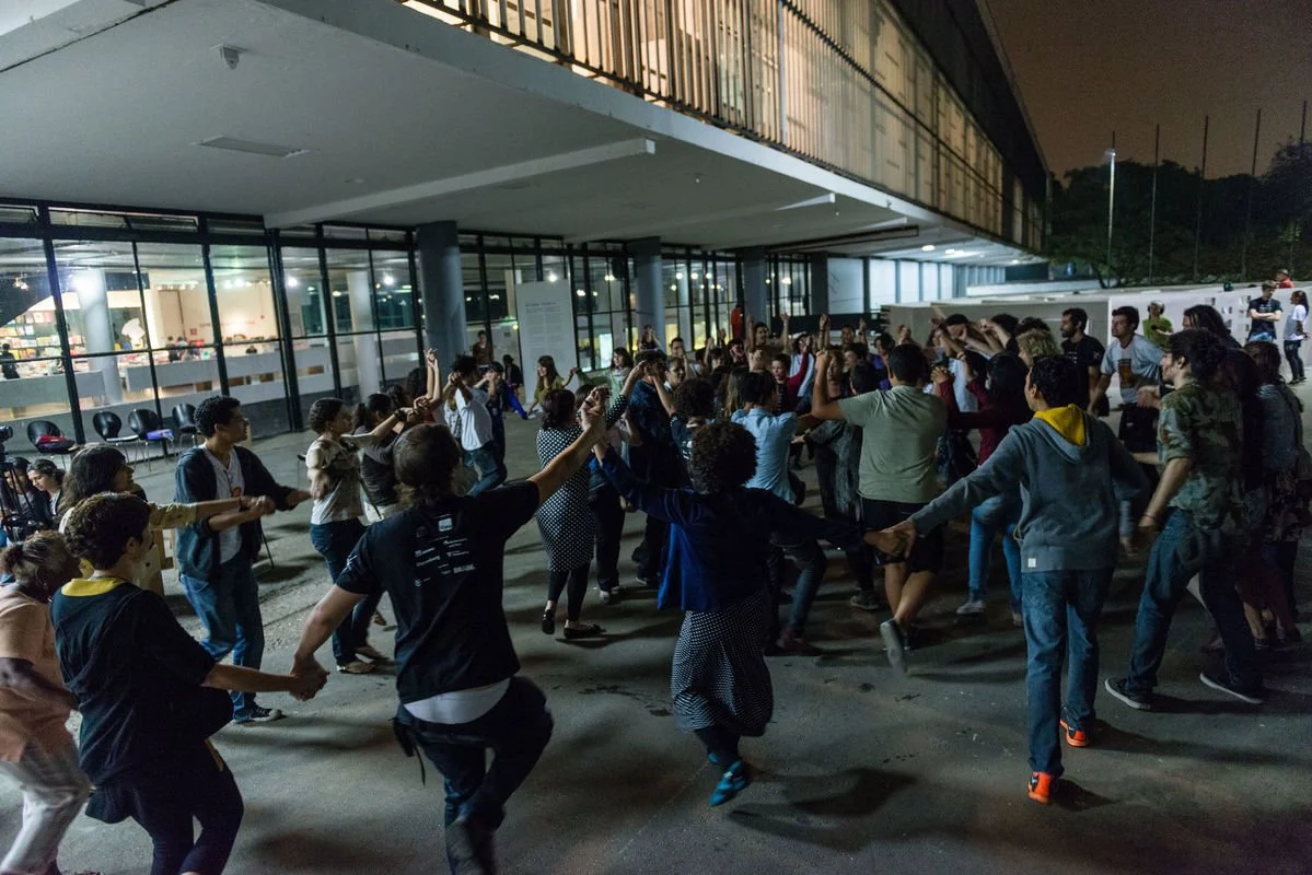Pessoas dançando ao ar livre à noite, formando um círculo, perto de um edifício de vidro.