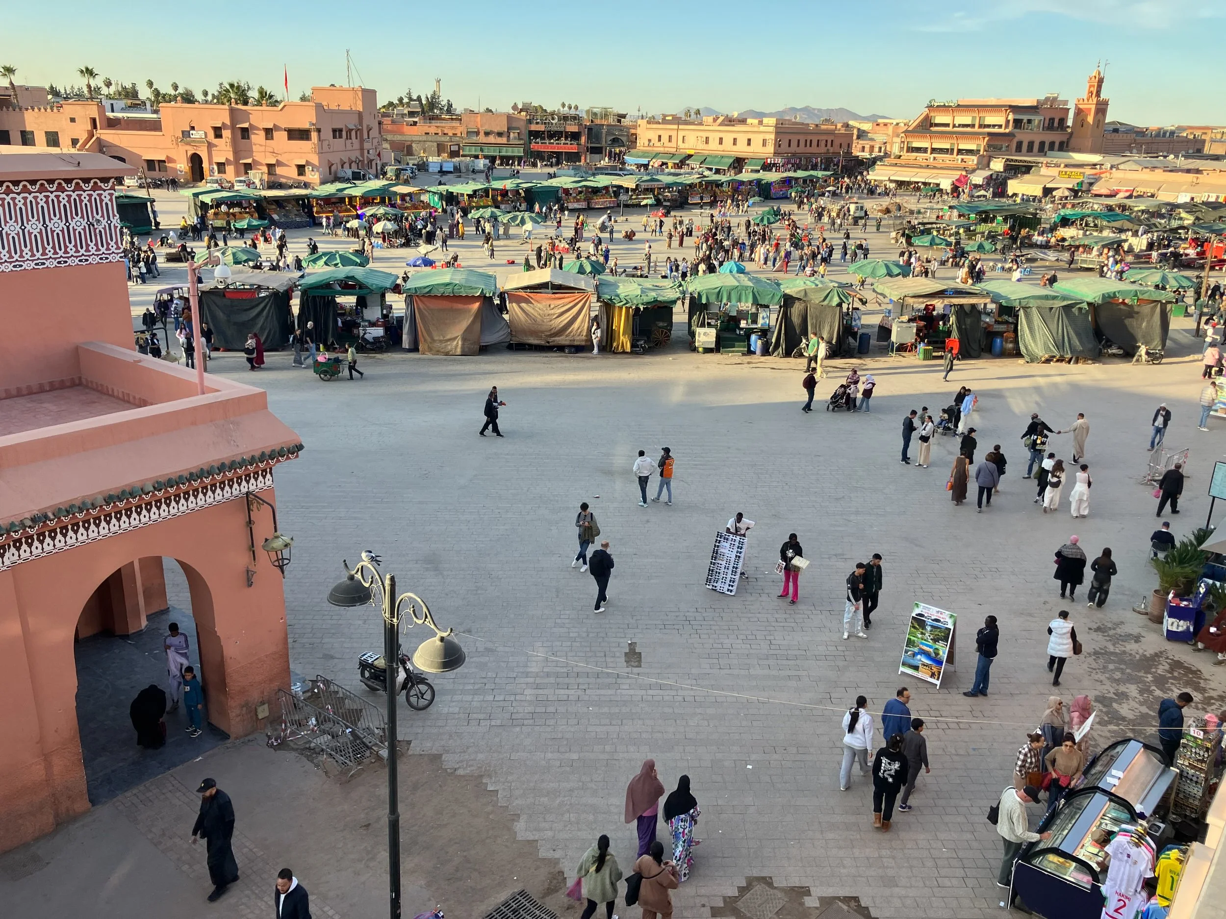 Praça movimentada com feirantes, vendedores em tendas, pessoas caminhando e comprando, além de lojas ao fundo e arquitetura marroquina.