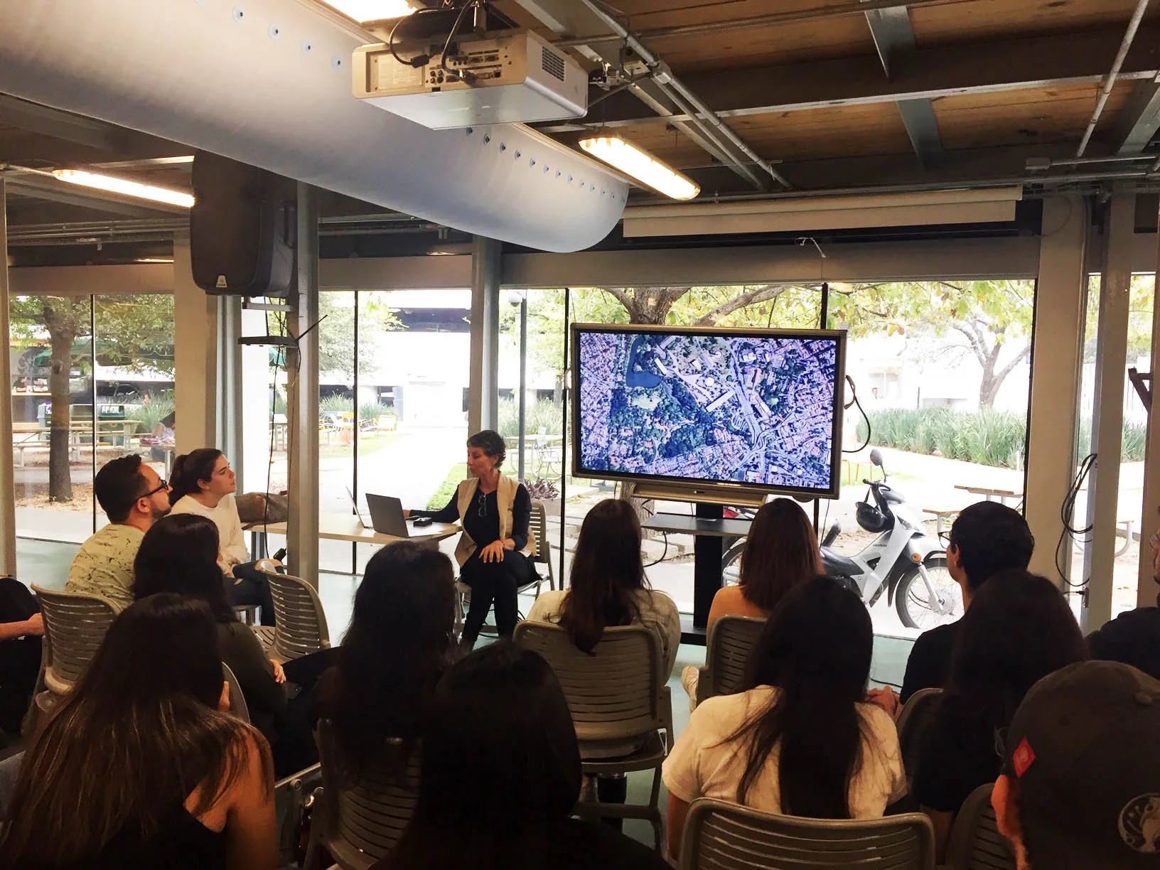 Palestrante apresentando para um grupo de pessoas em uma sala com visão para o exterior, incluindo árvores e uma motocicleta. Há uma tela grande exibindo uma imagem de satélite.