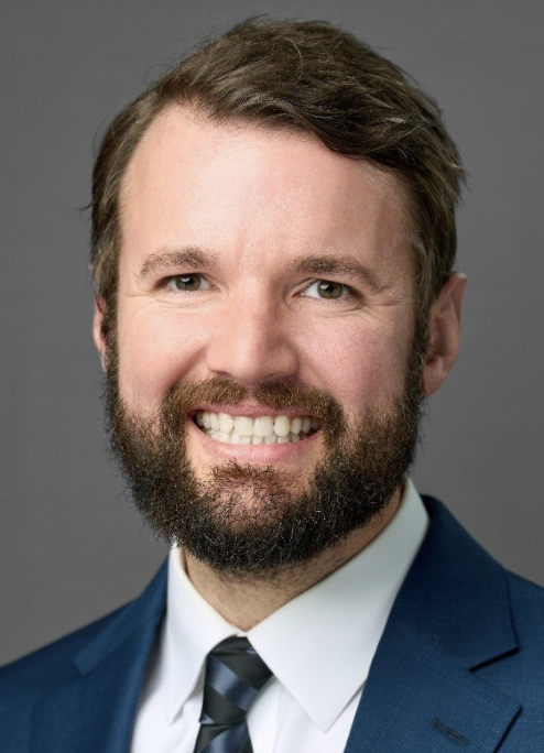 Headshot of a smiling man with a beard in a navy blue suit, white shirt, and striped tie against a gray background.