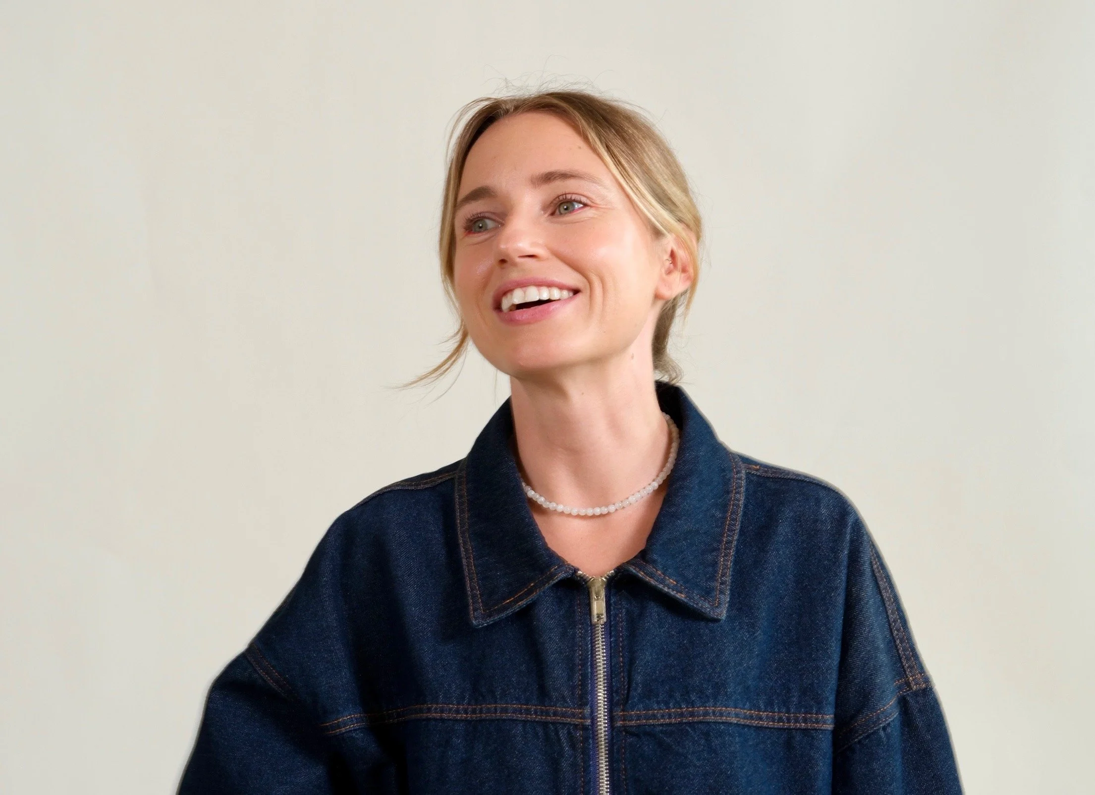 A smiling woman with blonde hair in a ponytail, wearing a denim jacket and a pearl necklace, standing against a light background.