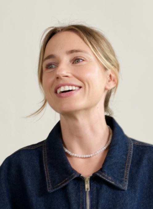 A smiling woman with blonde hair in a ponytail, wearing a denim jacket and a pearl necklace, standing against a light background.