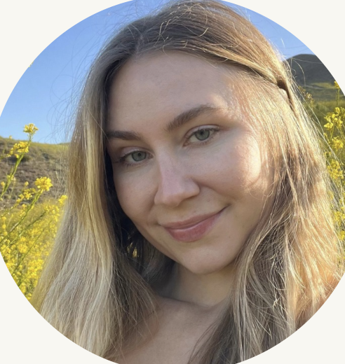 Close-up of a young woman with long blonde hair smiling outdoors with yellow flowers and a hill in the background.