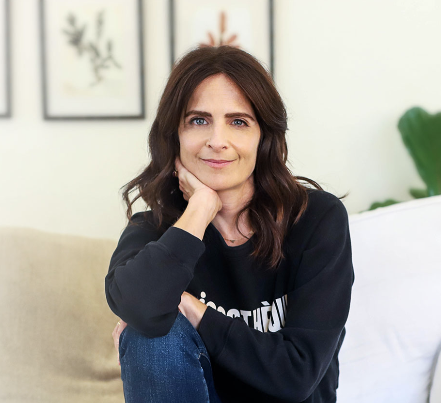 Woman with brown hair sitting on a white couch in a living room, smiling at the camera, with wall art and green plant in background.