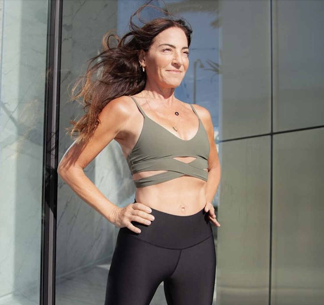A smiling woman with long, flowing brown hair stands with her hands on her hips in front of a modern glass building. She is wearing a green sports bra with cut-out details and black workout leggings.