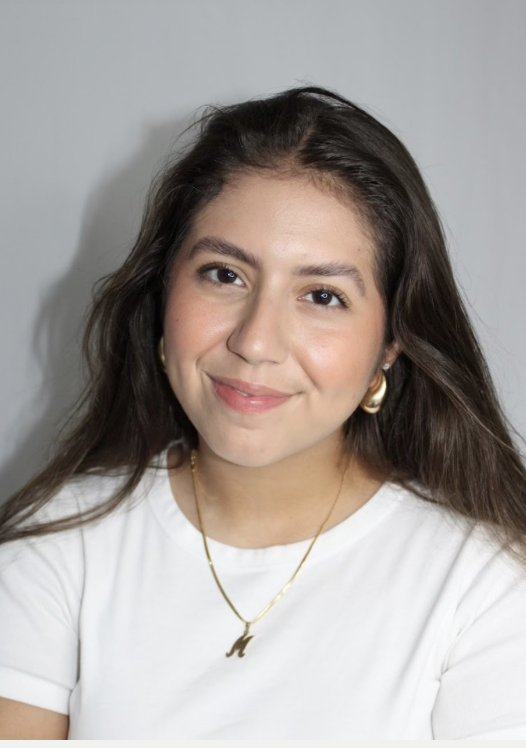 A young woman with long brown hair smiling at the camera, wearing a white T-shirt, gold earrings, a gold necklace, and a small gold pendant.