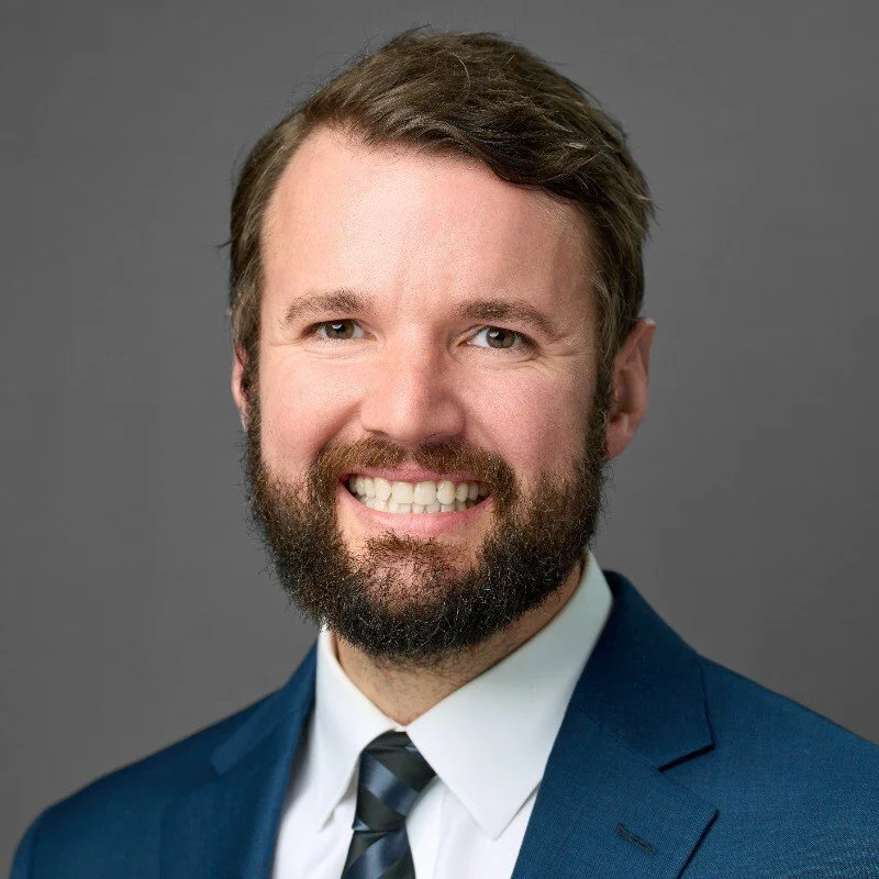 Headshot of a smiling man with a beard in a navy blue suit, white shirt, and striped tie against a gray background.