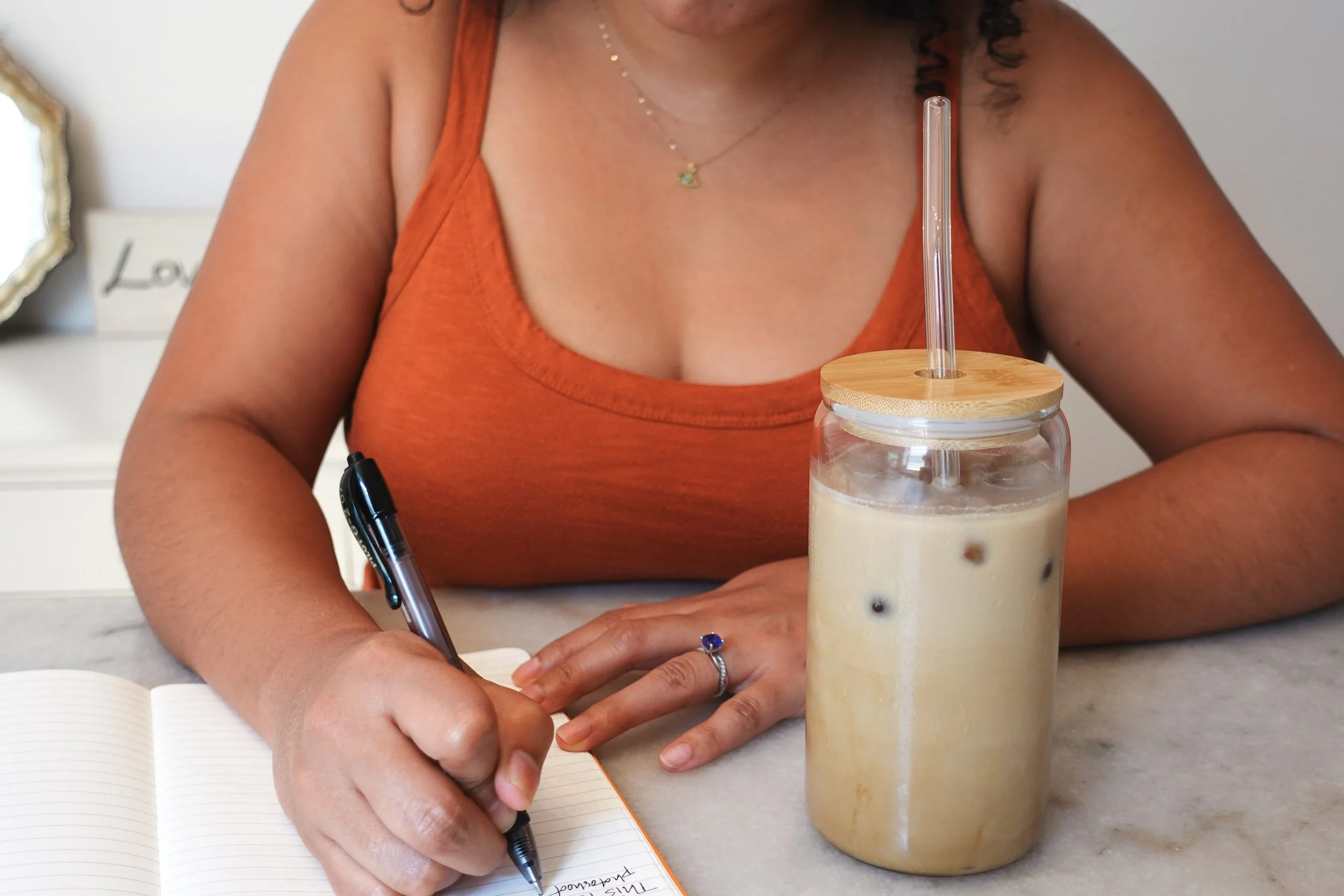 Close up of a hand writing in a notebook next to a cup of iced coffee.