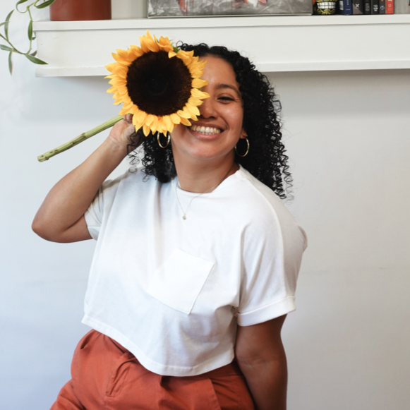 Book Coach Sabrina smiles at the camera while holding a sunflower over one of her eyes.
