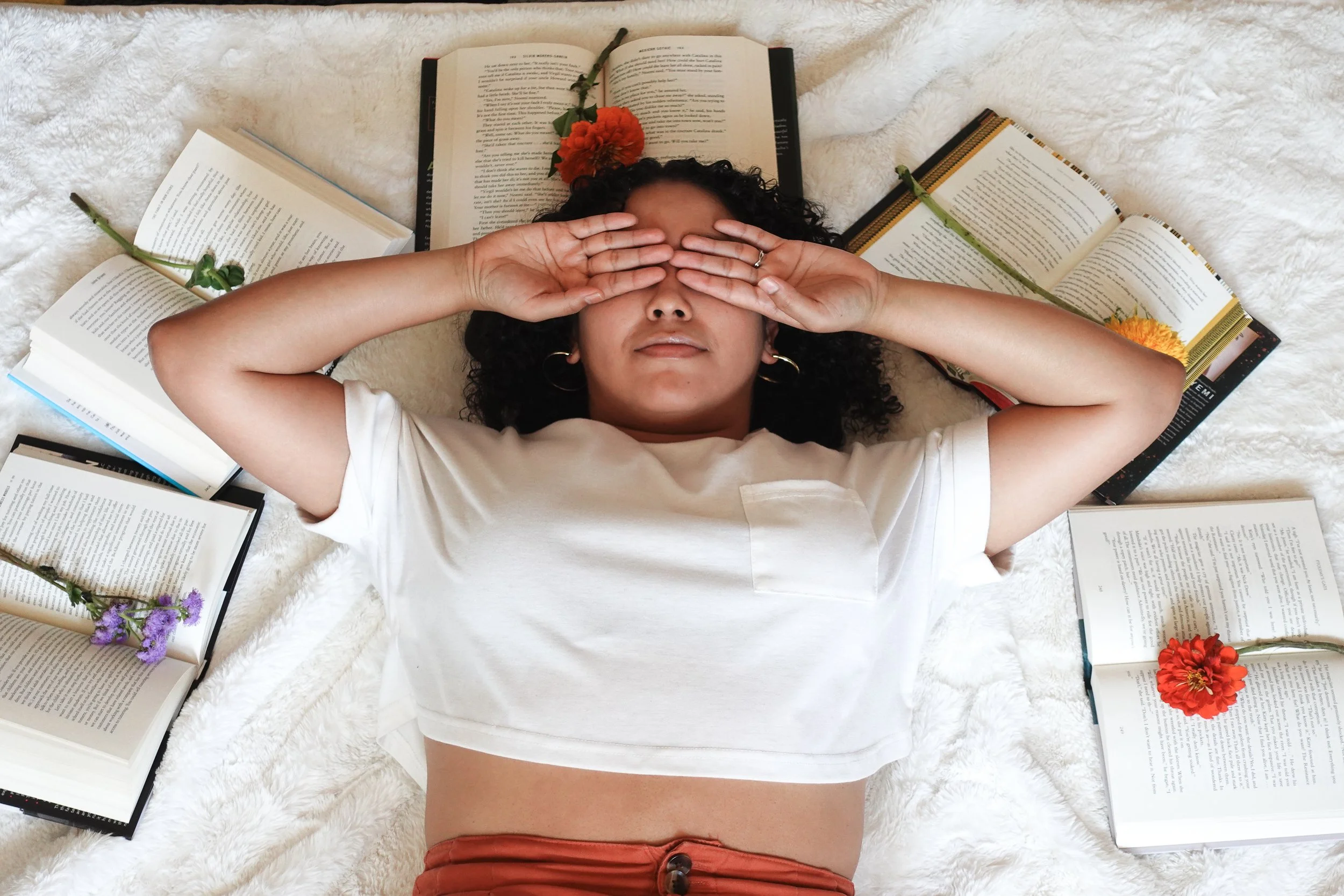 Book coach Sabrina lies on a white rug with her hands over her eyes and a semicircle of books around her with flowers on the pages.