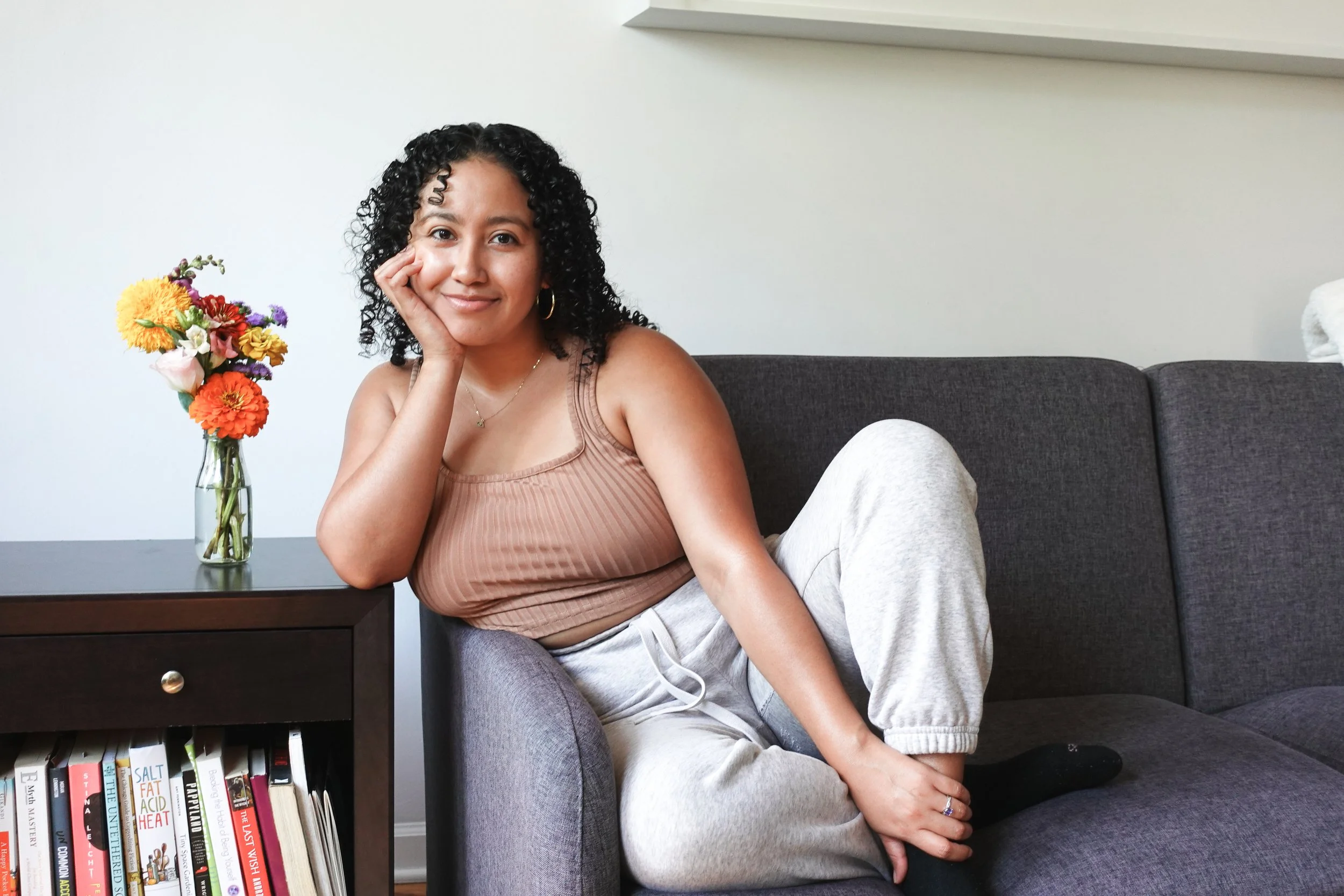 Book coach Sabrina sits on a couch next to a side table with flowers on it and smiles softly at the camera.