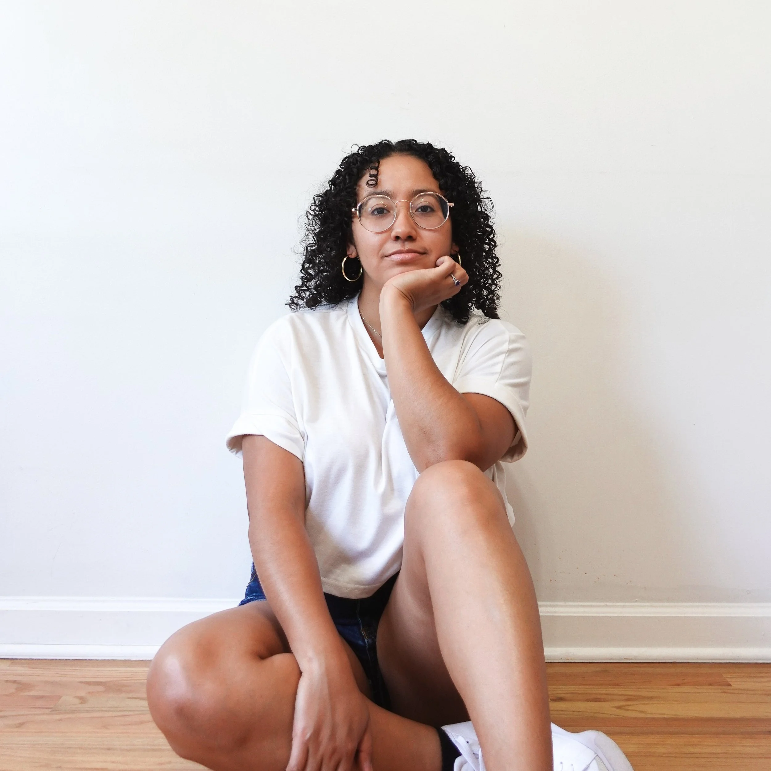 Book coach Sabrina sits on the floor in front of a blank white wall and looks at the camera with neutral expression.