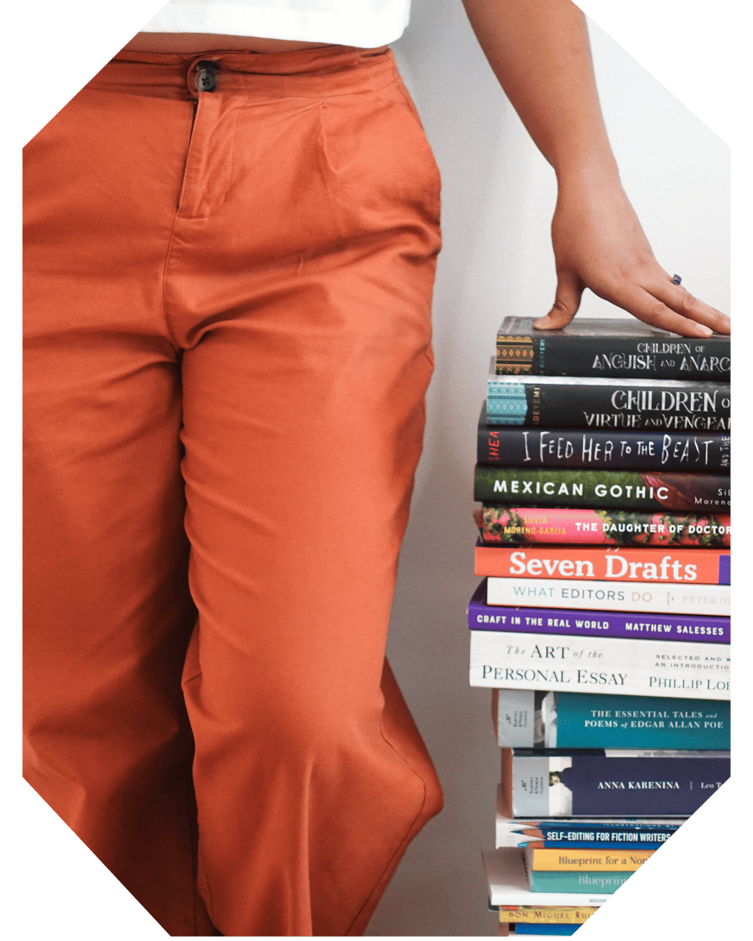 Book Coach Sabrina stands next to a bookstack that reaches her hips and puts one hand on top of the books. Only her legs, her hand, and the books are visible.