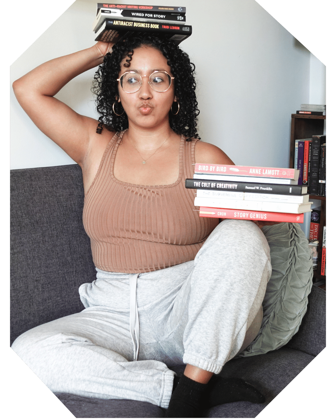 Book Coach Sabrina sits on the couch and balances one stack of book on her knee and another stack on her head and she looks off camera and purses her lips.