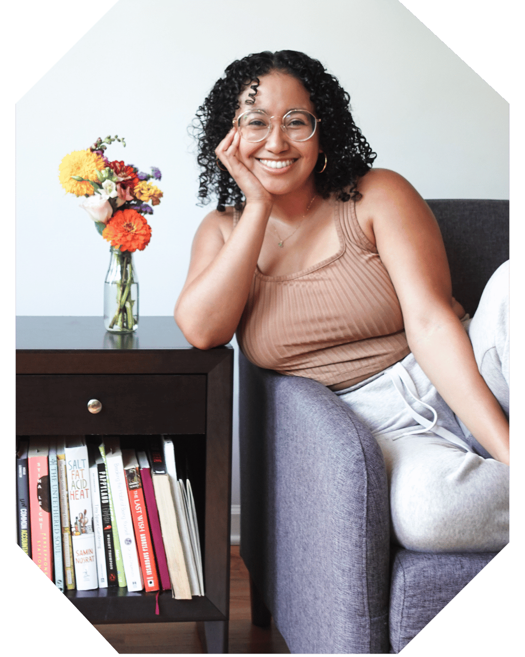 Book Coach Sabrina sits on a couch and smiles at the camera while leaning on a side table with flowers on top and books below.