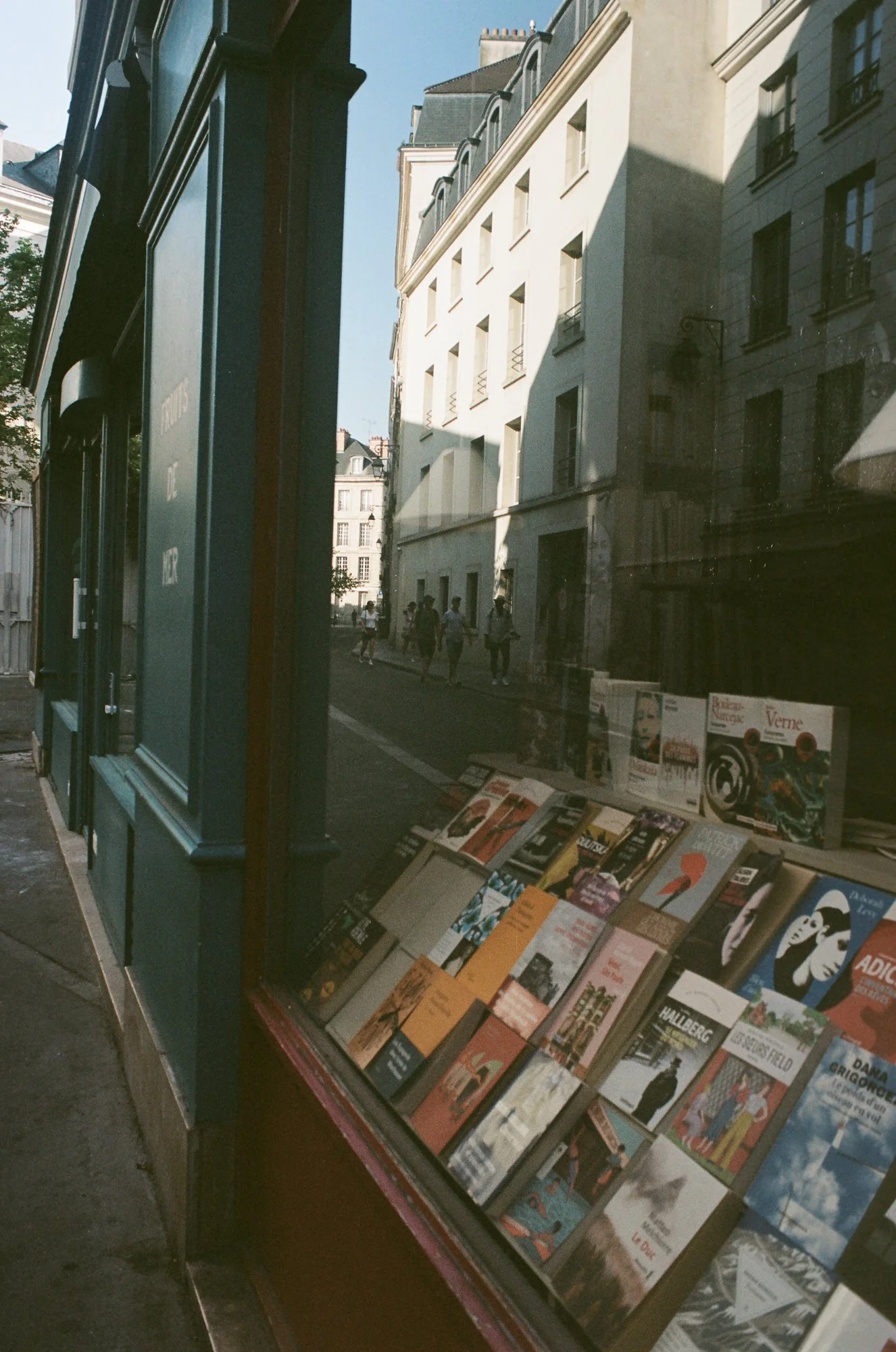 A street view with a green bookstore window display of various books, reflecting buildings and people walking on the sidewalk outside.
