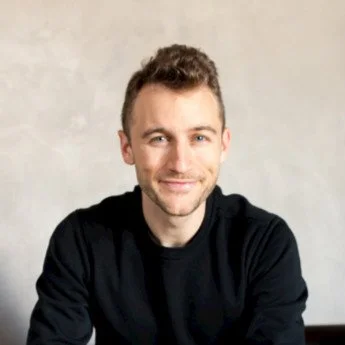 A young man with short, curly hair and a beard, smiling while wearing a black shirt against a light-colored background.