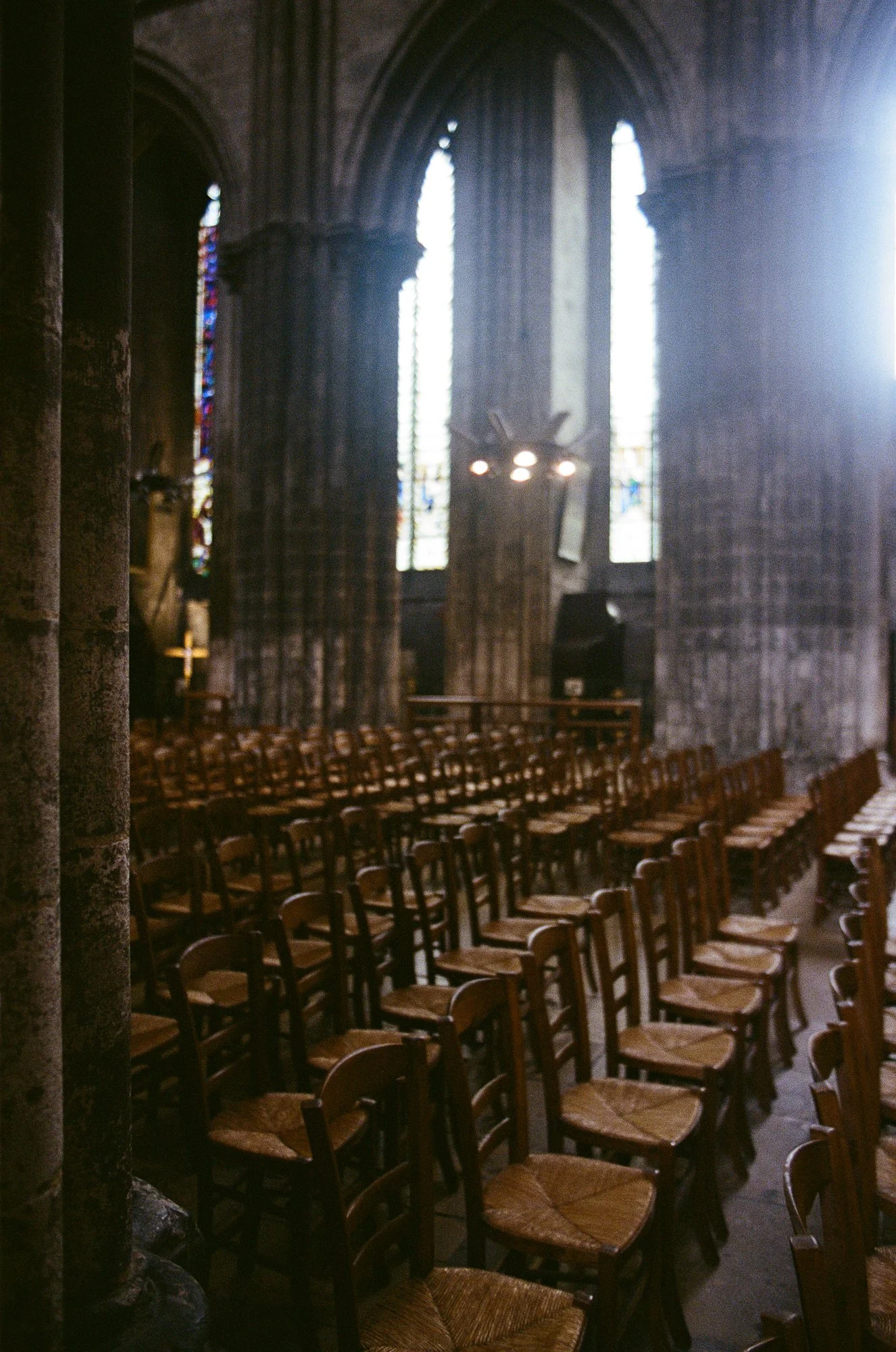 Interior of a church with wooden chairs arranged in rows, stained glass windows, and stone walls. Sunlight streams in from the right side.