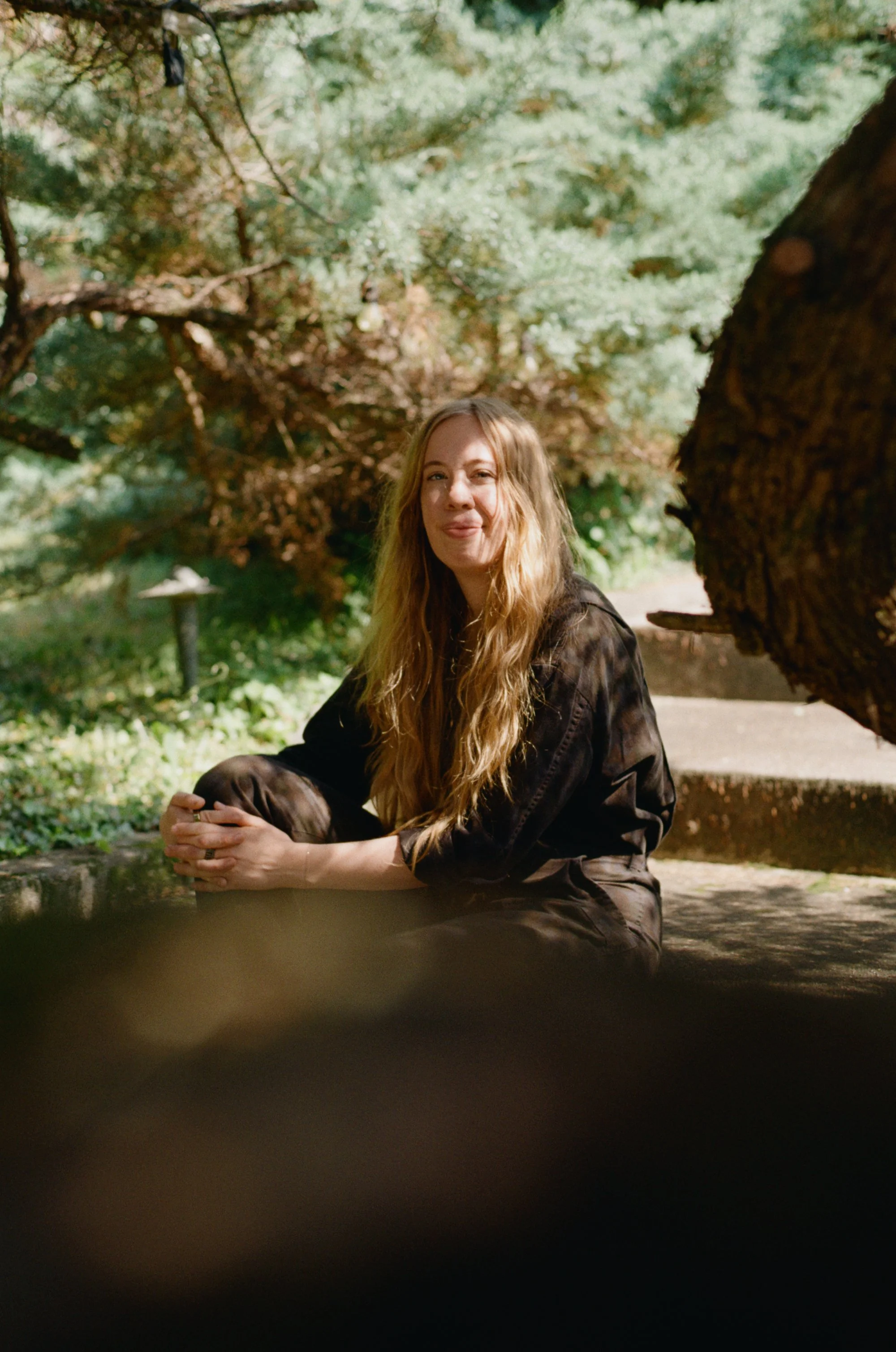 A woman with long blonde hair sitting outdoors on a stone surface, surrounded by greenery and trees, making a playful face.