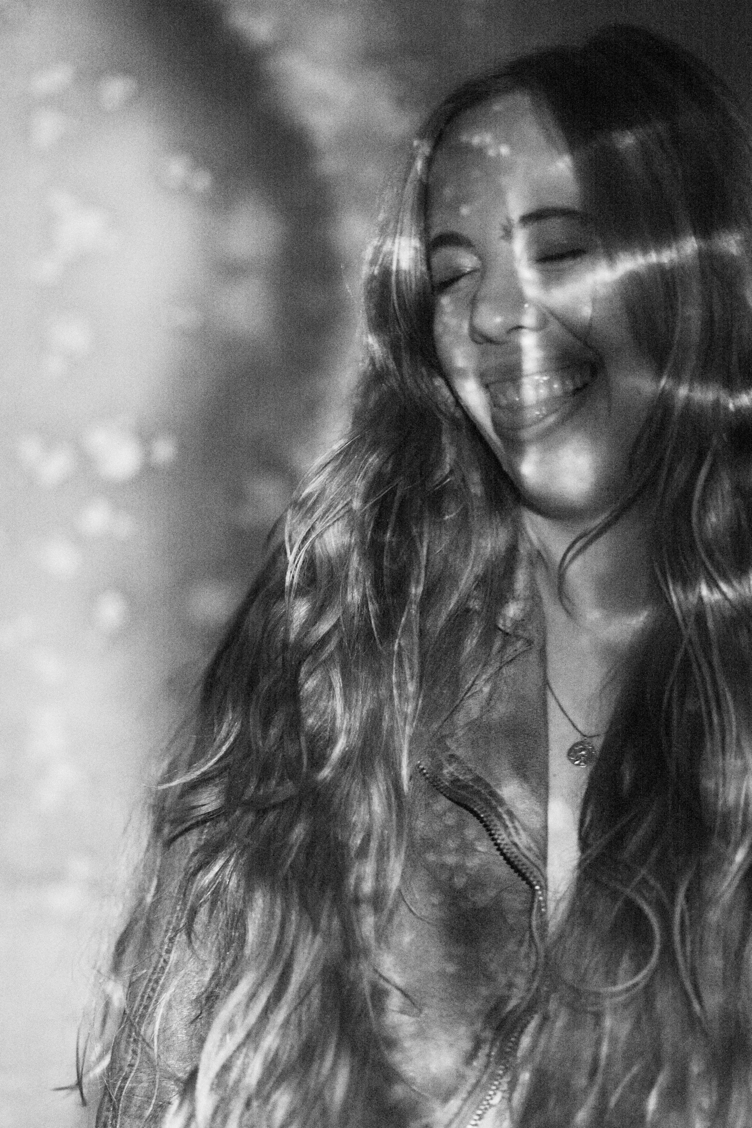 A black and white photo of a young woman with long wavy hair, smiling with eyes closed, wearing a jacket with a zipper and a necklace.