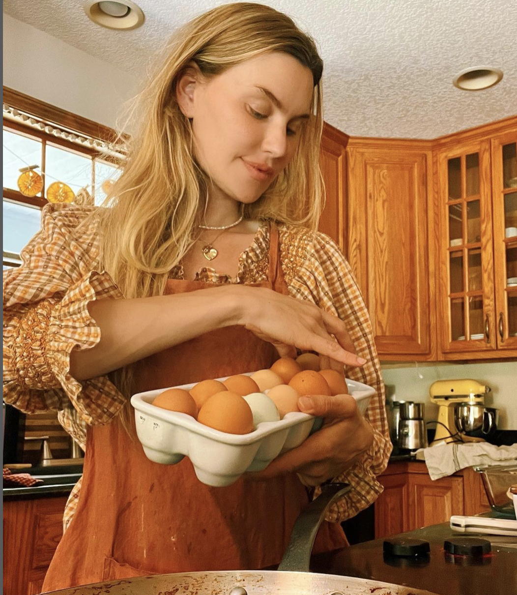 A woman in a kitchen holding a carton of brown and white eggs while cooking. She is wearing an orange apron and a checkered shirt, looking down at the eggs.