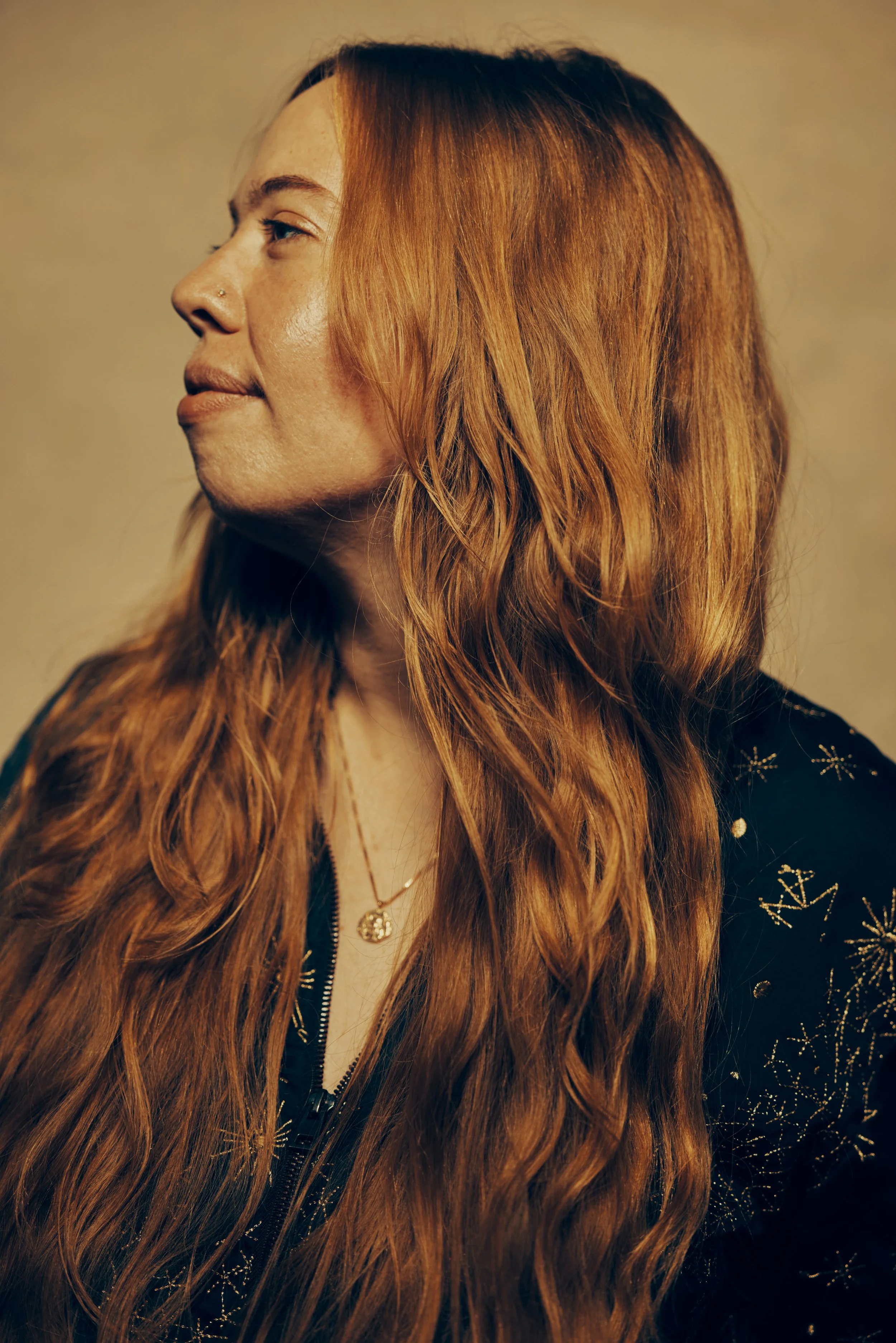 A woman with long, wavy red hair in profile view looking to the left, wearing a black jacket with sparkly star designs and a gold necklace.