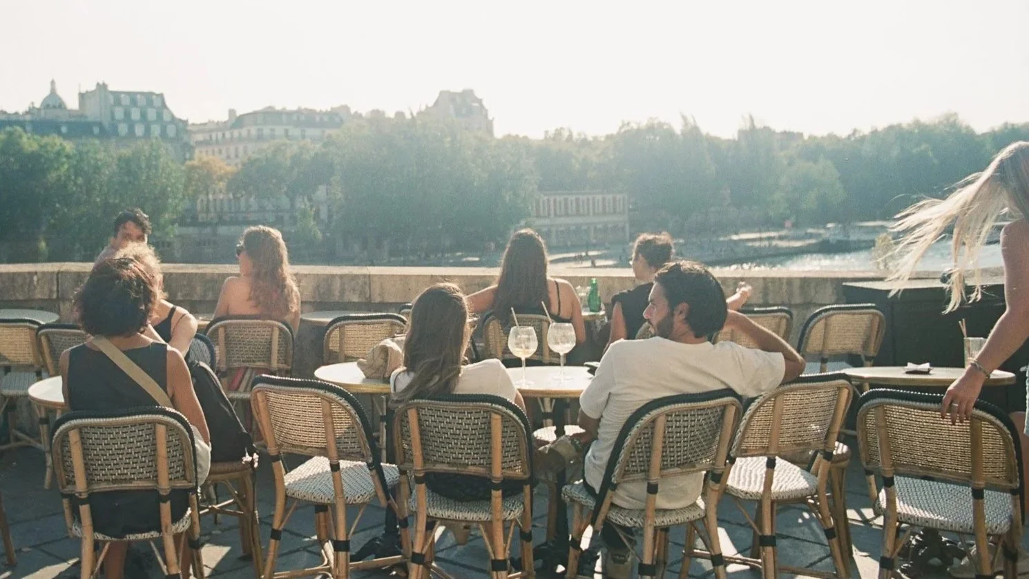 Group of people sitting on outdoor terrace, overlooking river and city skyline in the background, during daytime.