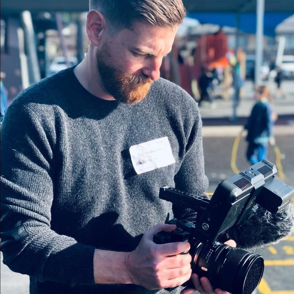 Man with beard and gray sweater holding professional video camera outdoors, with street scene and people in background.
