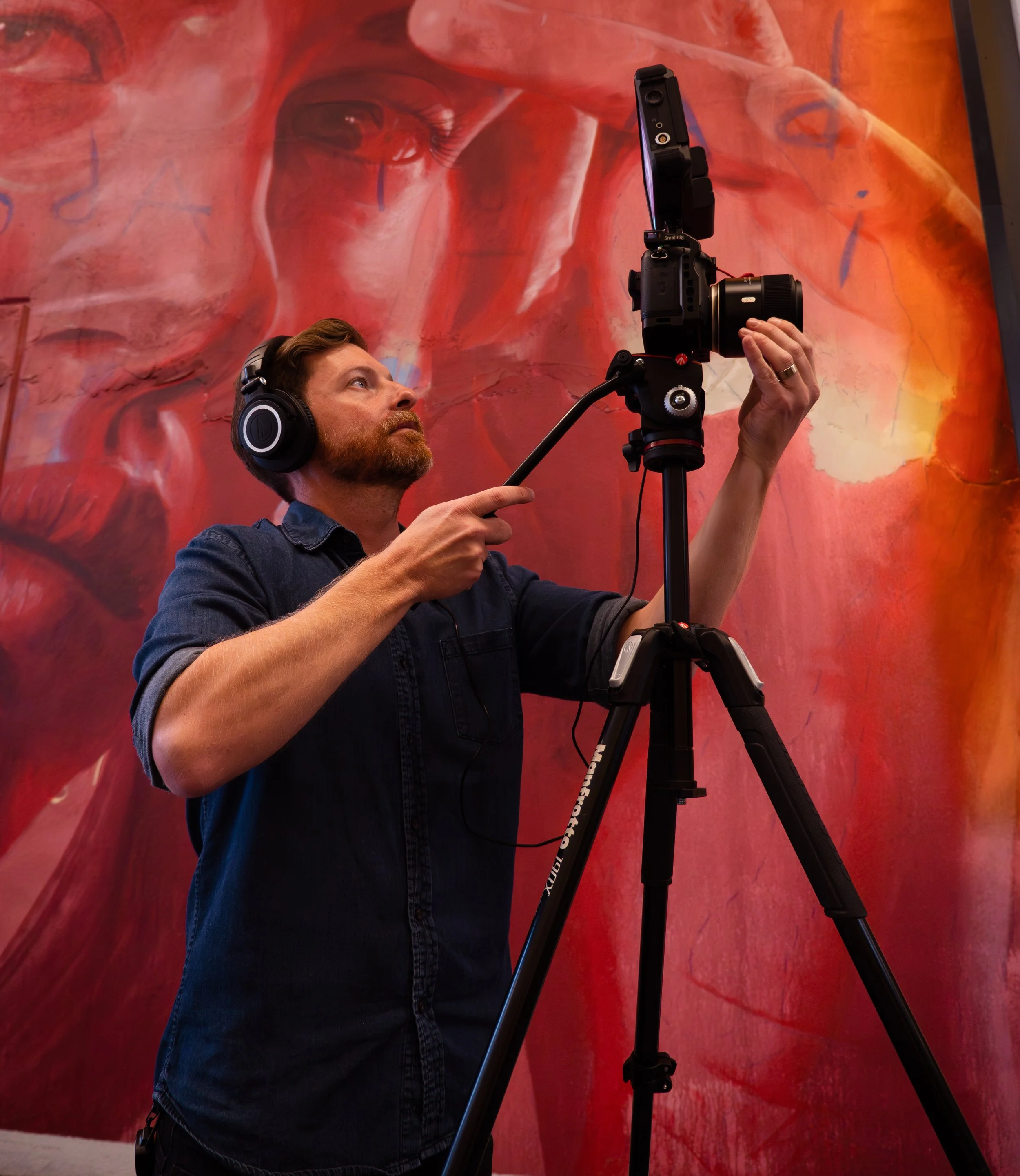A man wearing headphones adjusting a camera on a tripod in front of a large abstract red and orange painting.