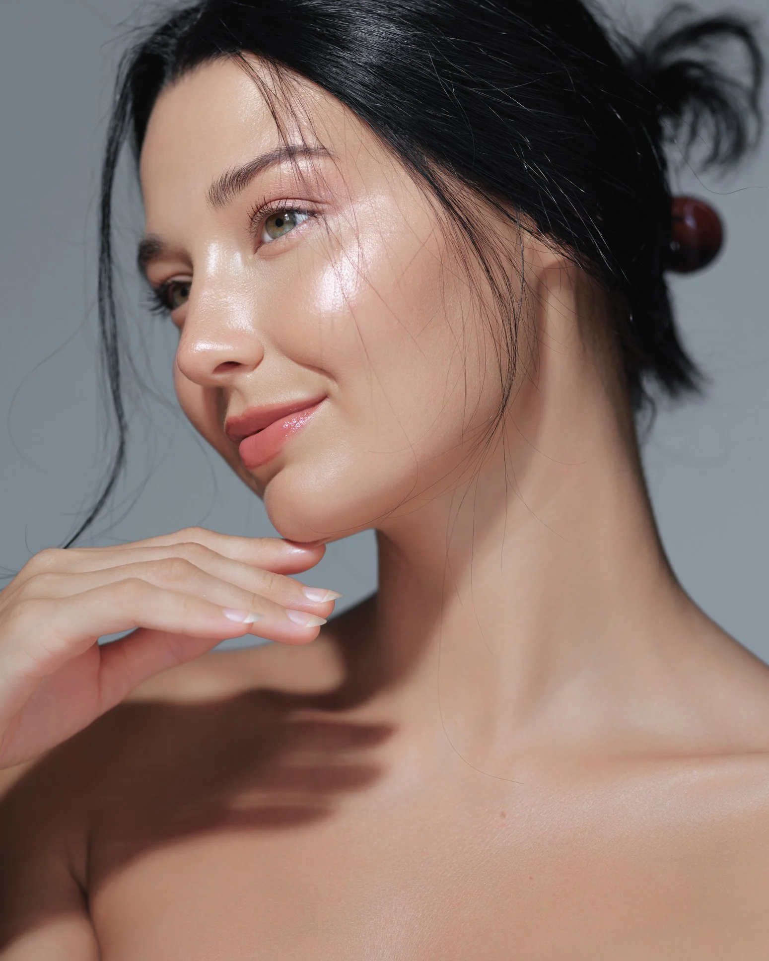 Close-up of a woman with flawless skin, light makeup, and dark hair styled with hairpins, touching her chin with her hand against a neutral background.
