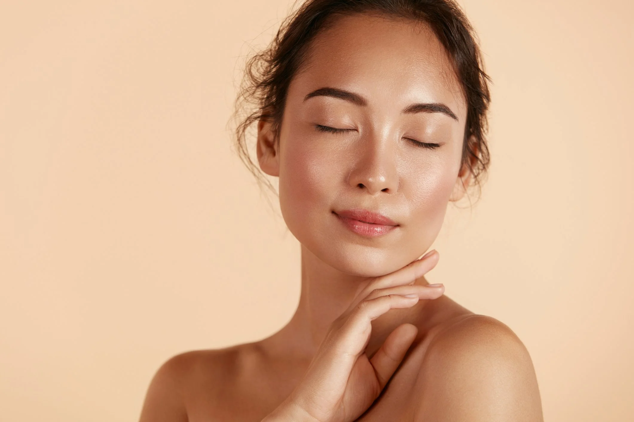 Close-up of a woman with clear, glowing skin, eyes closed, smiling slightly, touching her chin with her hand, against a light beige background.
