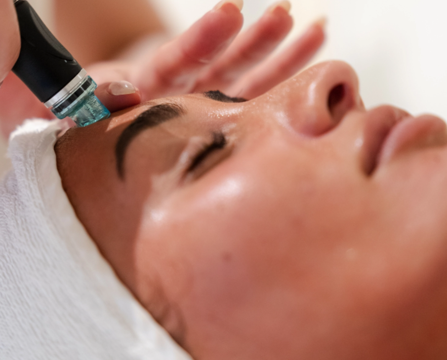 A person is receiving a facial treatment with a HydraFacial device, lying down with eyes closed, while a technician wearing gloves holds the equipment near their face.