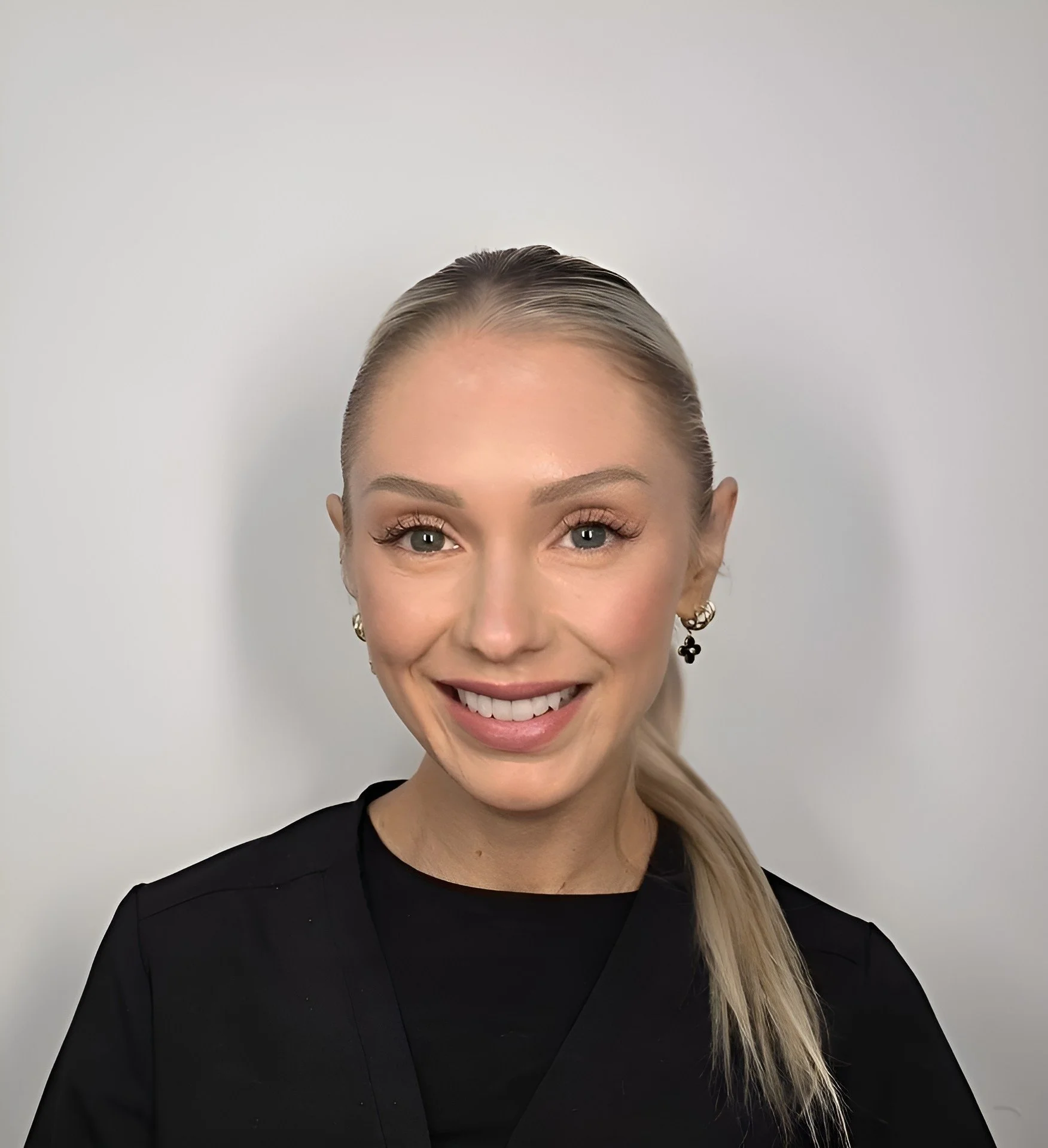 A smiling woman with blonde hair in a ponytail, wearing hoop earrings with black cross charms and a black top, standing against a plain light-colored background.