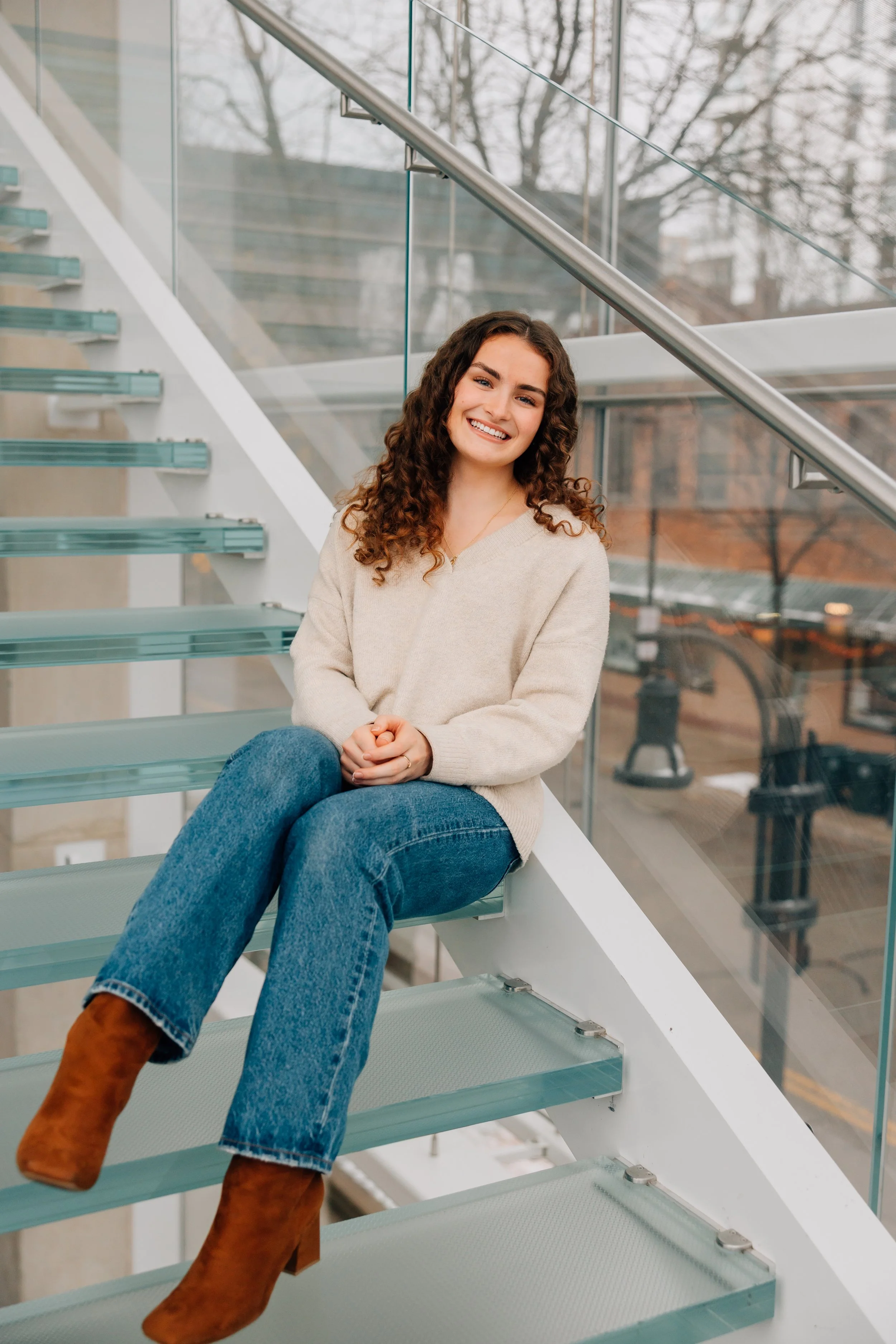 A woman with curly brown hair sitting on glass stairs, smiling, wearing a beige sweater, blue jeans, and brown boots, in a modern building with large windows.